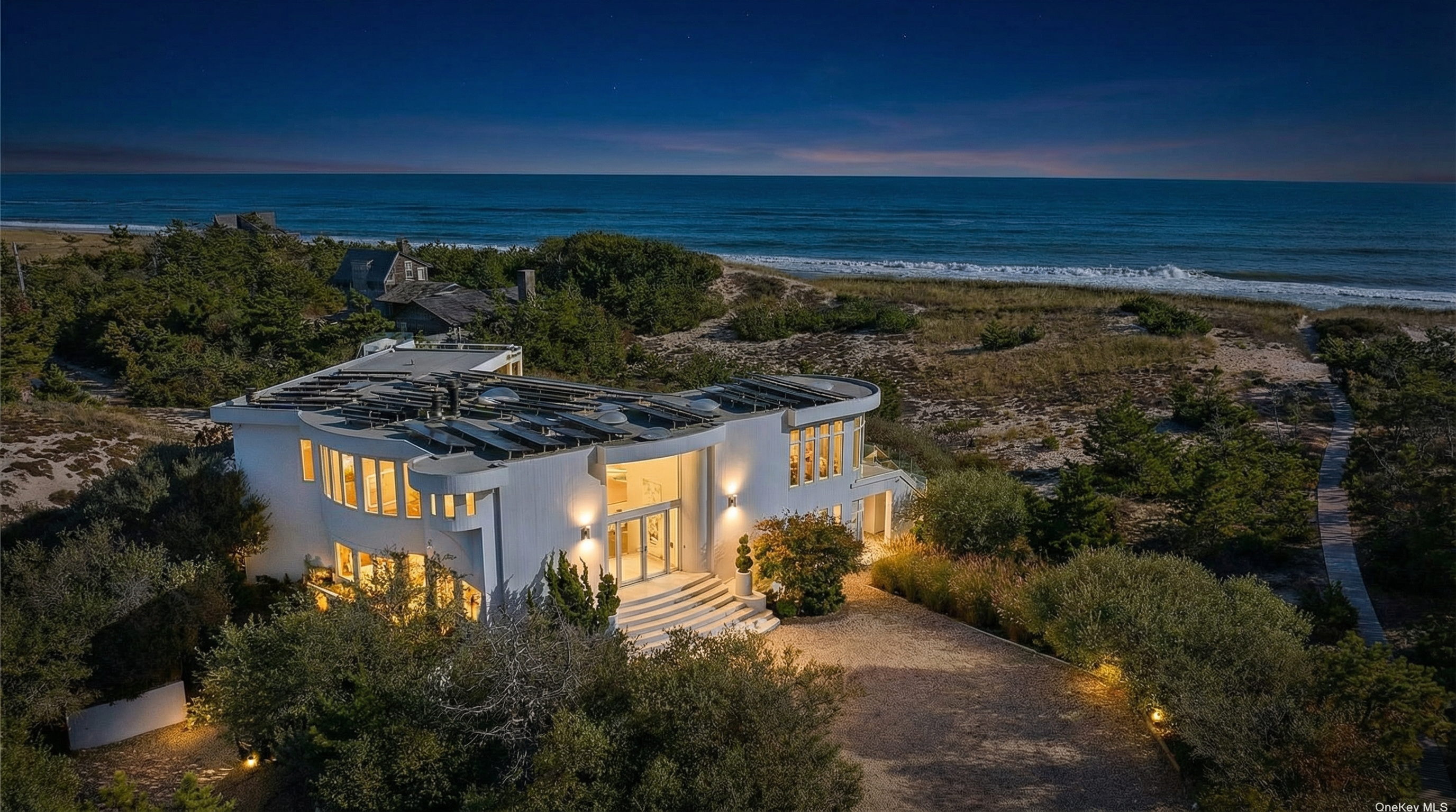 This aerial view showcases a modern, white beach house with a curved design and multiple solar panels on the roof. The house is set amidst lush greenery and sandy dunes, with ocean views visible in the background. A long driveway leads up to the house, adding to the property's secluded and private feel.