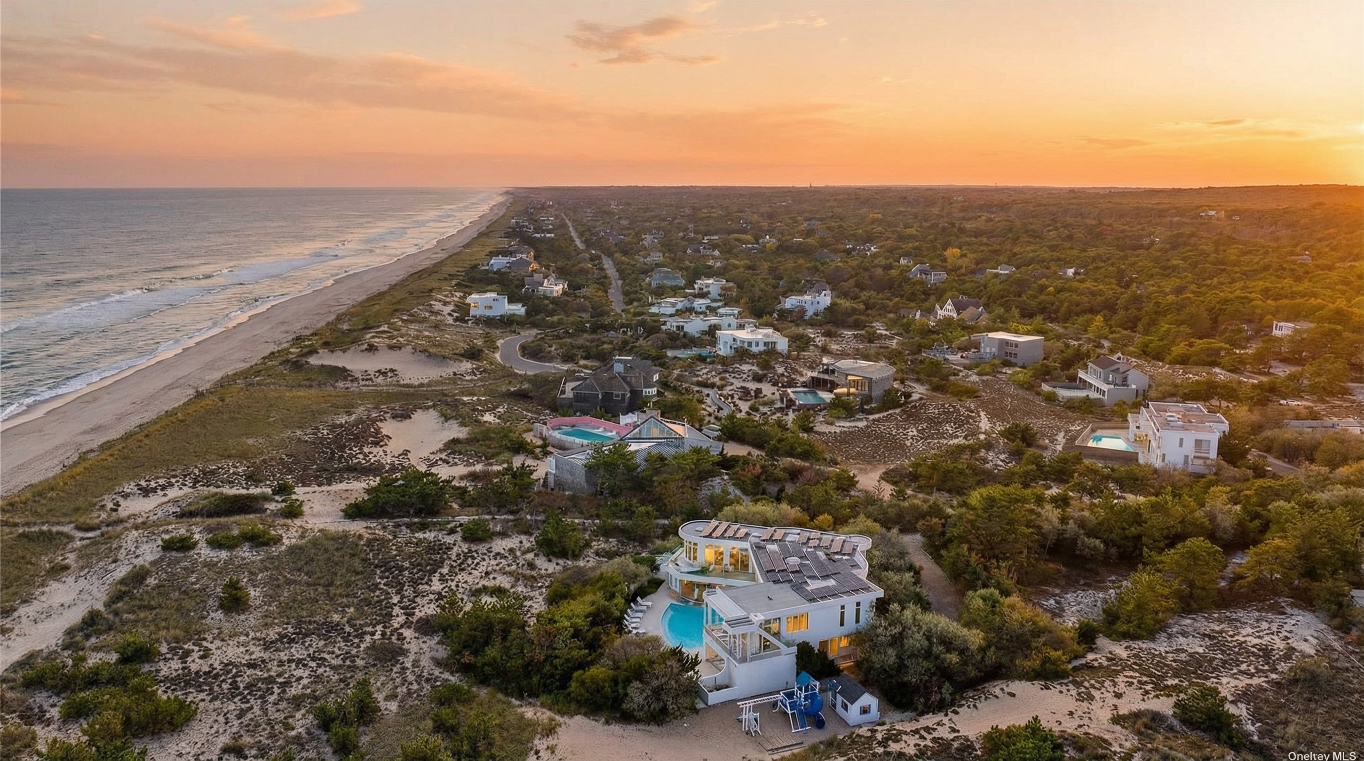 This aerial shot showcases a luxury beachfront property with various architectural details. The property features a modern design with a swimming pool, solar panels, and a playground. The landscape includes lush greenery, sand dunes, and a view of the ocean, creating an inviting atmosphere.