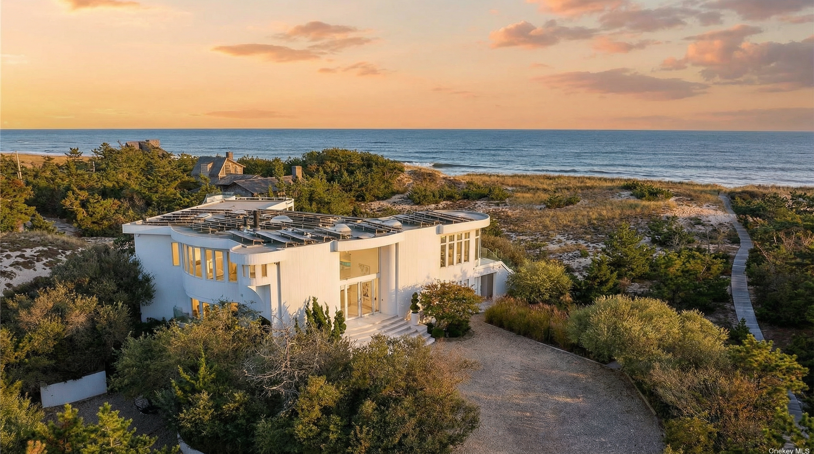This aerial view showcases a stunning modern beachfront home. The house features a white exterior with a flat roof adorned with solar panels. The property includes a well-maintained driveway, lush landscaping, and direct access to the beach via a wooden walkway, emphasizing privacy and luxury.
