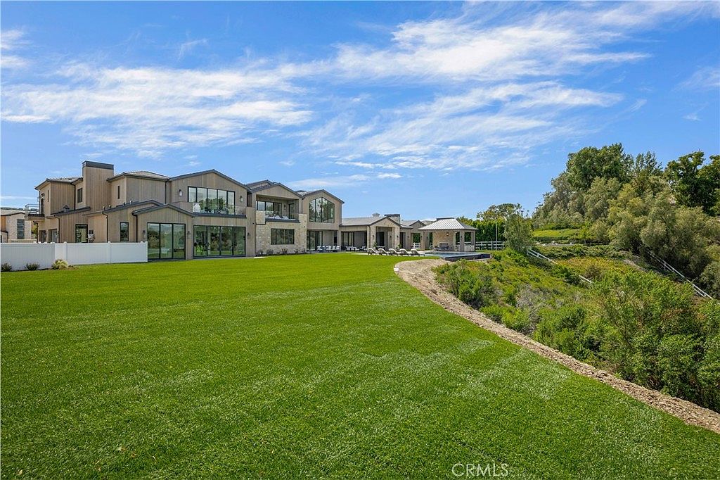 This expansive rear view of a luxury estate showcases a modern architectural design with large glass windows and doors that seamlessly connect the indoor living spaces to the outdoors. A vast, manicured green lawn stretches across the foreground, leading toward a private pool area and a covered patio structure. The property is situated on a hillside, offering a sense of privacy and scenic views of the surrounding natural landscape.