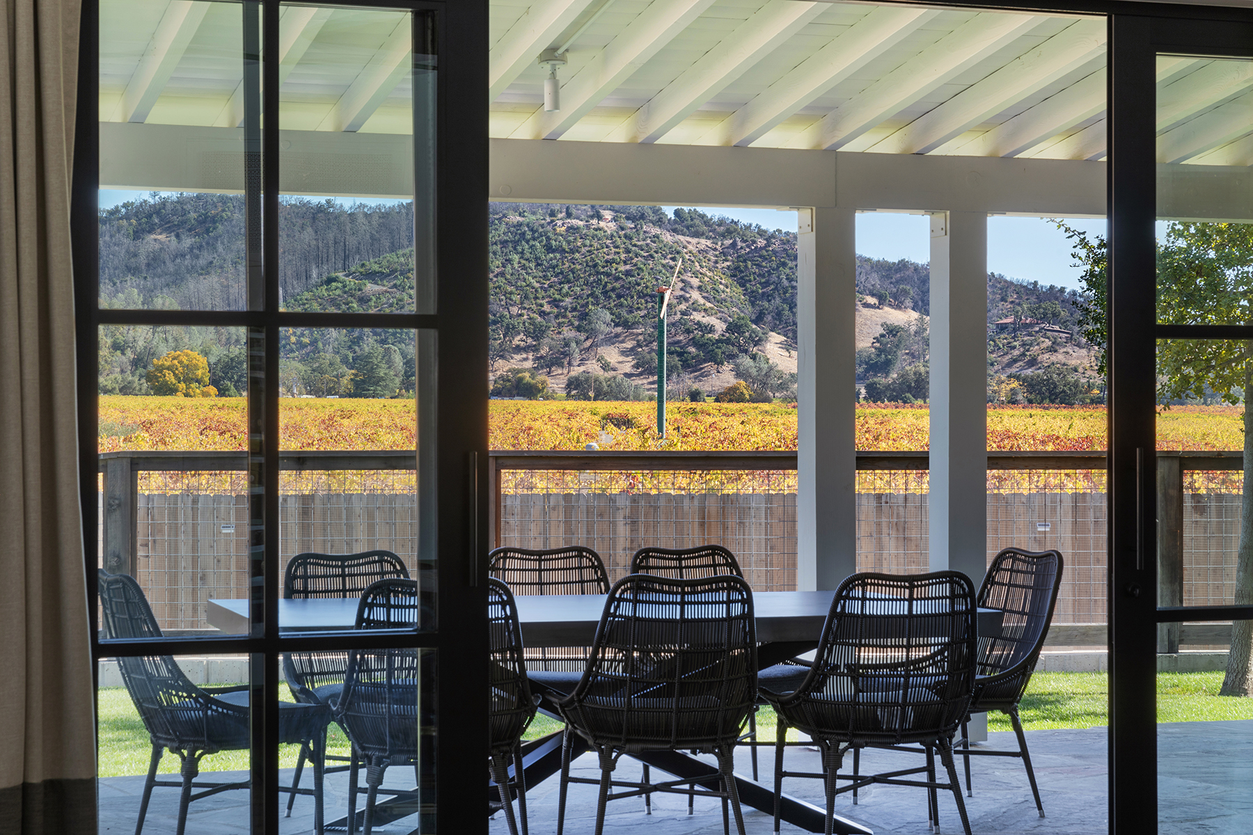 This covered outdoor patio features a contemporary dining set with dark wicker chairs and a sleek table, overlooking a scenic vineyard and hilly landscape. The area is framed by elegant black-framed glass doors, creating a seamless transition between indoor and outdoor living spaces. The property is highlighted by its serene, natural setting and refined, modern design.