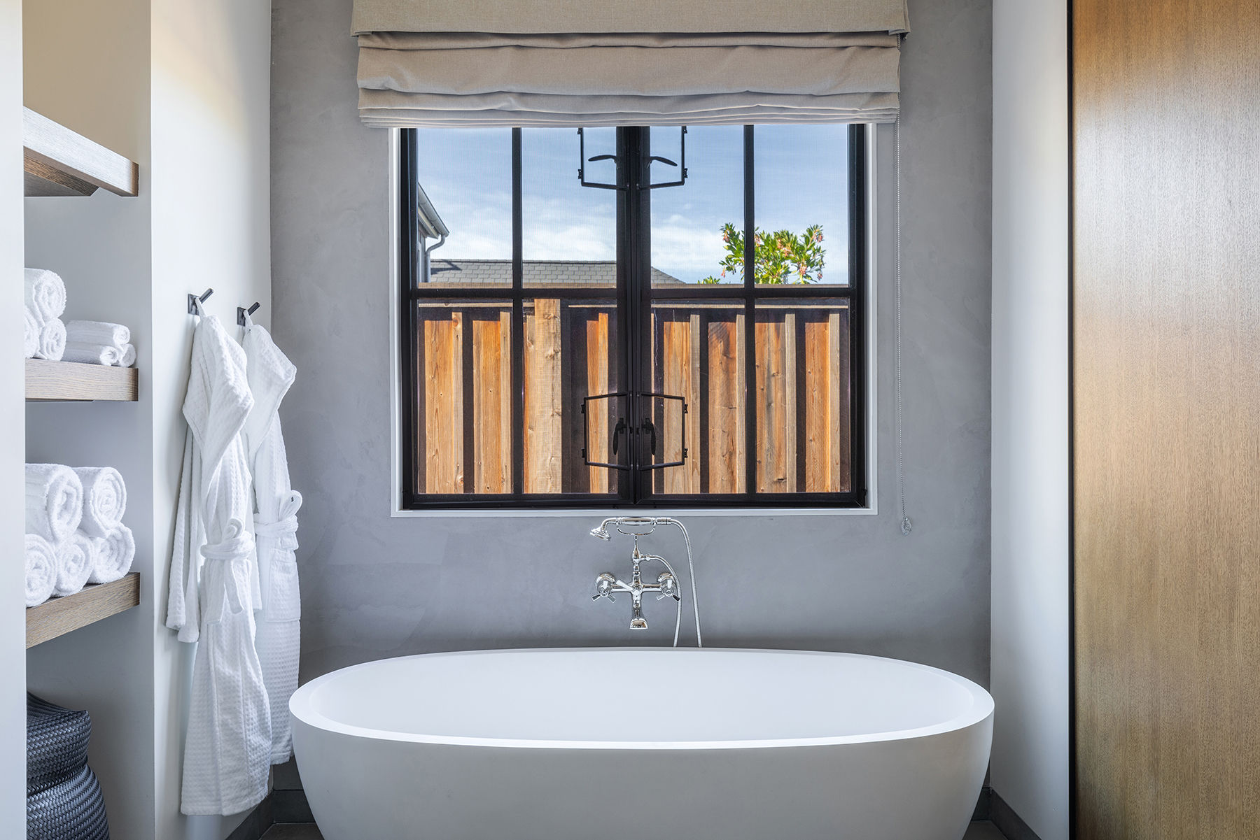This serene primary bathroom features a deep, white freestanding soaking tub centered beneath a multi-pane window. The space is accented by a textured gray wall, floating wood shelving stocked with plush white towels, and high-end chrome plumbing fixtures. Two white bathrobes hang neatly on the wall, enhancing the spa-like and luxurious aesthetic of the room.