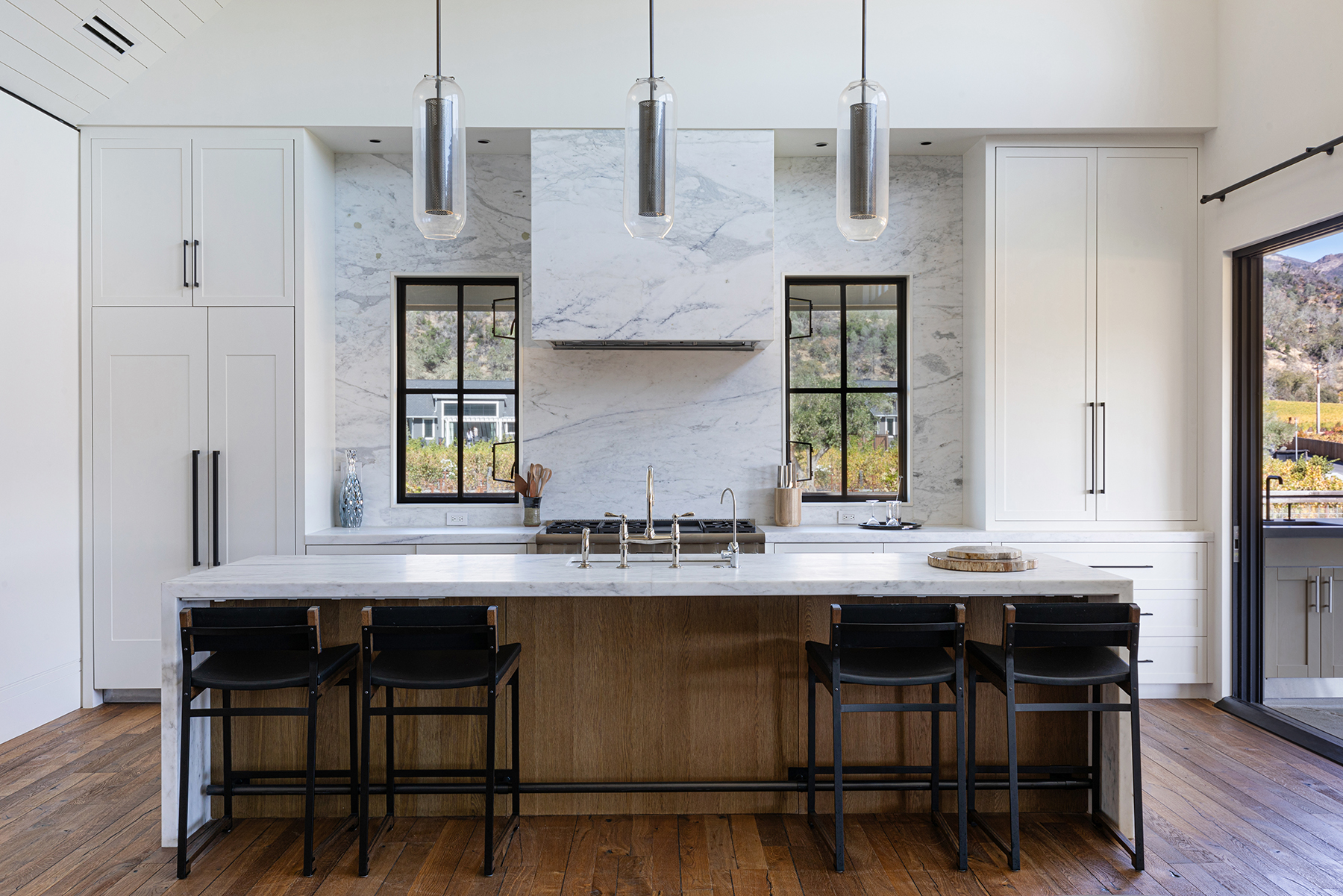 This stunning contemporary kitchen features a substantial marble-topped island with seating for four, complemented by custom white cabinetry and a matching marble backsplash. The space is anchored by a chef-grade range hood and elegant pendant lighting, creating a bright and sophisticated atmosphere. With large windows flanking the stove, the kitchen offers beautiful views and a seamless connection to the outdoors.