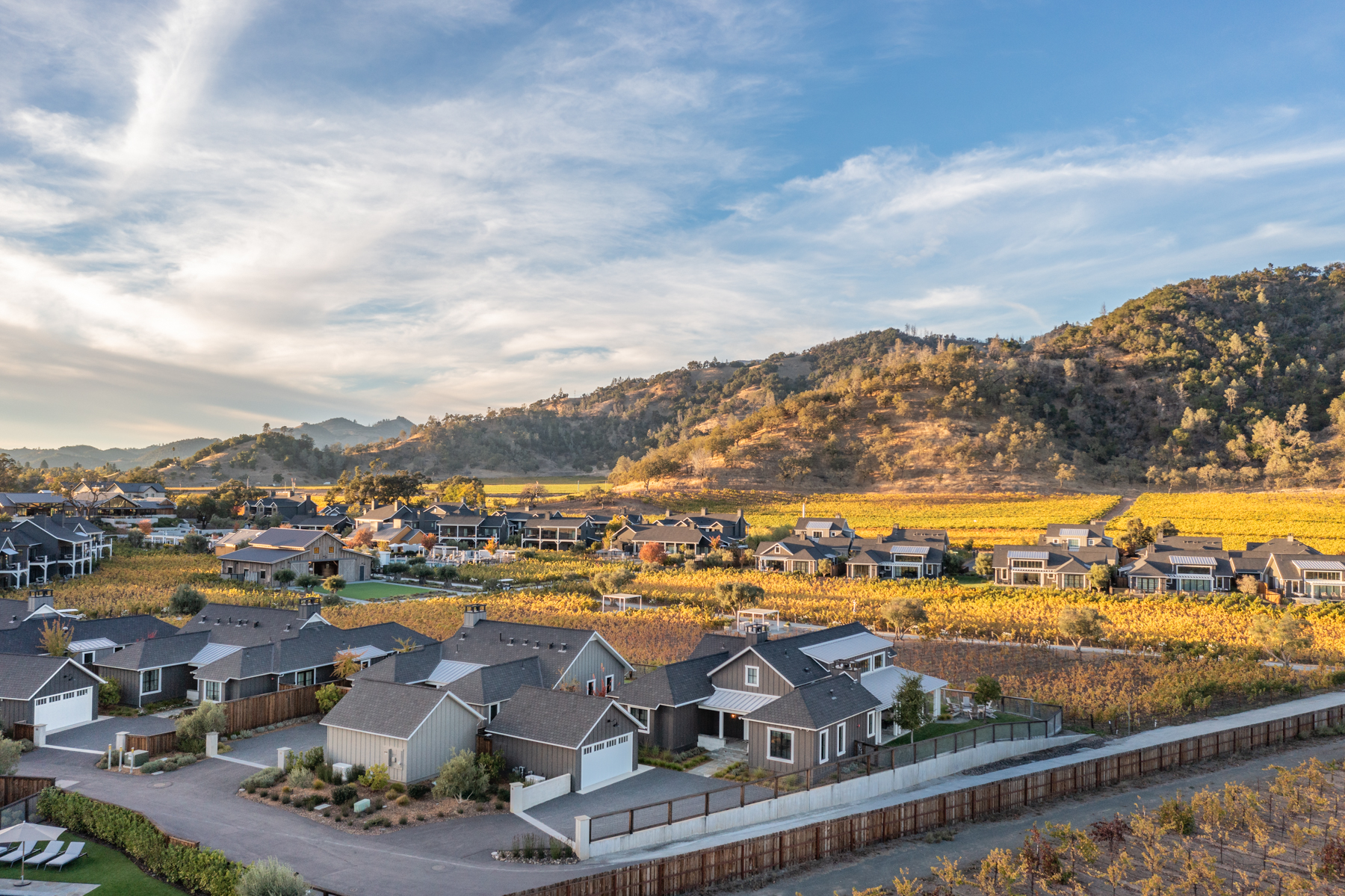 This panoramic aerial view captures an upscale residential community nestled amidst vast, golden-hued vineyards at sunset. The development features modern farmhouse-style homes with dark grey siding and pitched roofs, well-integrated into the scenic, rolling landscape of Northern California. The peaceful, manicured neighborhood combines rustic vineyard charm with refined architectural design, offering a serene luxury living environment.