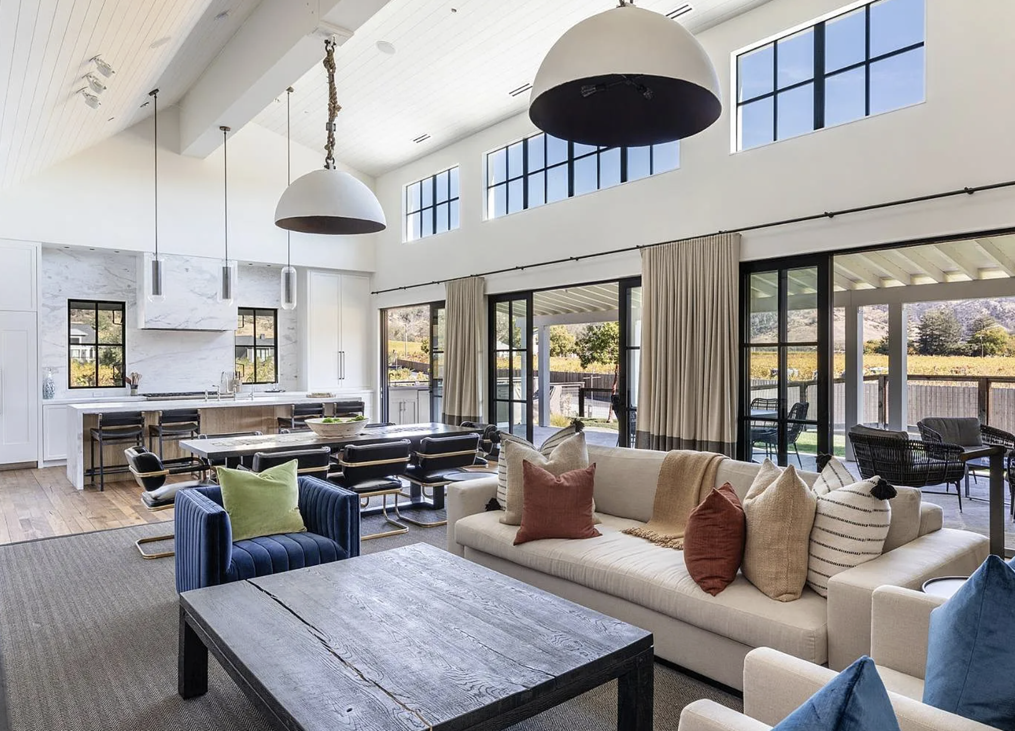 This spacious open-plan living and dining area features vaulted, beam-accented ceilings and abundant natural light provided by expansive floor-to-ceiling windows and sliding glass doors. The interior design blends a sophisticated, modern style with a neutral palette, highlighted by a navy velvet armchair, a substantial rustic wooden coffee table, and a marble kitchen island. The seamless connection to the outdoor patio and scenic vineyard views creates a bright, luxurious, and inviting atmosphere ideal for entertaining.