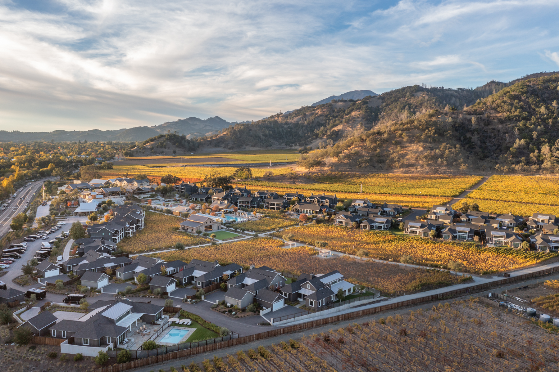 This high-angle, aerial perspective showcases an upscale residential community seamlessly integrated into a expansive vineyard landscape. The homes feature modern farmhouse architecture with dark roof pitches and organized layouts, highlighting the scenic transition from meticulously manicured living spaces to the sprawling, golden-hued autumn vines framed by rugged mountains.