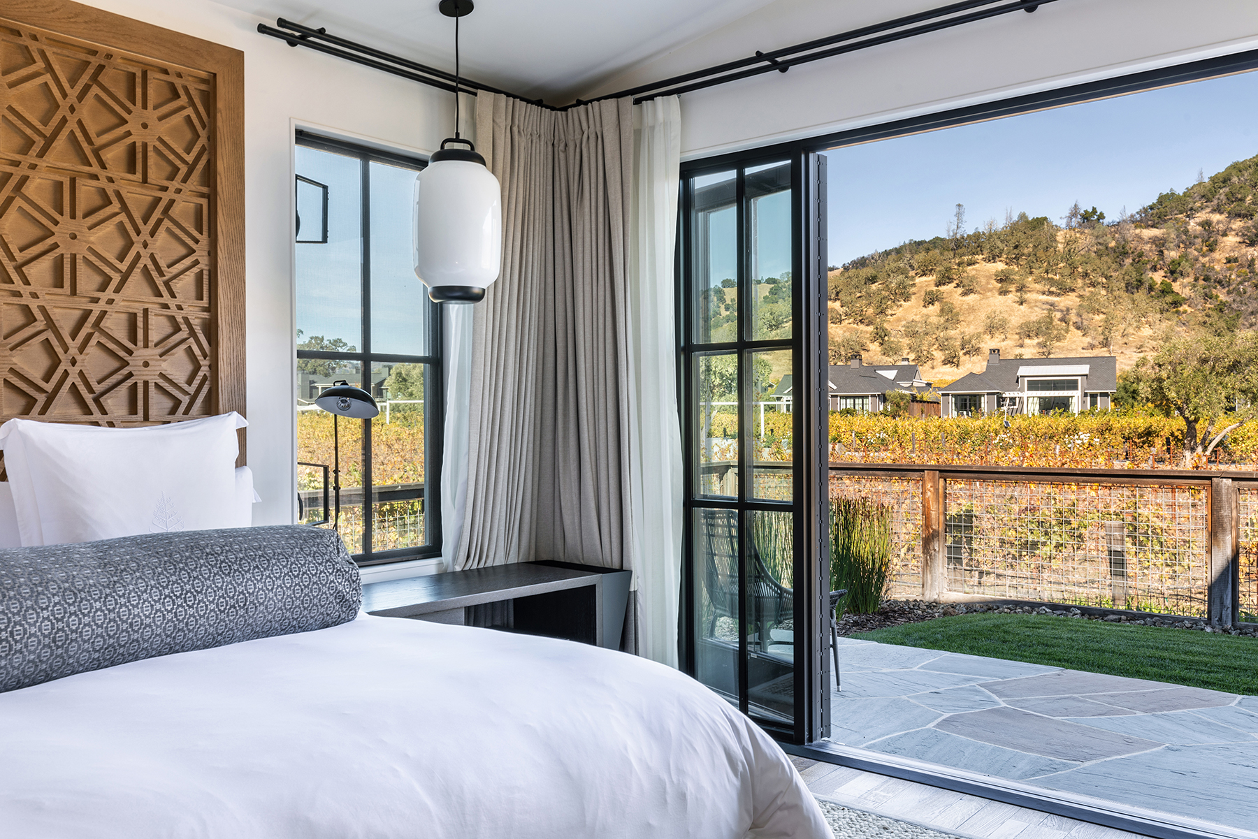 This sophisticated primary bedroom showcases a striking custom geometric wood headboard and minimalist decor. The room seamlessly blends indoor and outdoor living with floor-to-ceiling glass doors that open onto a private stone patio overlooking a scenic vineyard. High-end finishes, such as the lantern-style pendant light and elegant drapery, contribute to a luxurious and tranquil atmosphere.