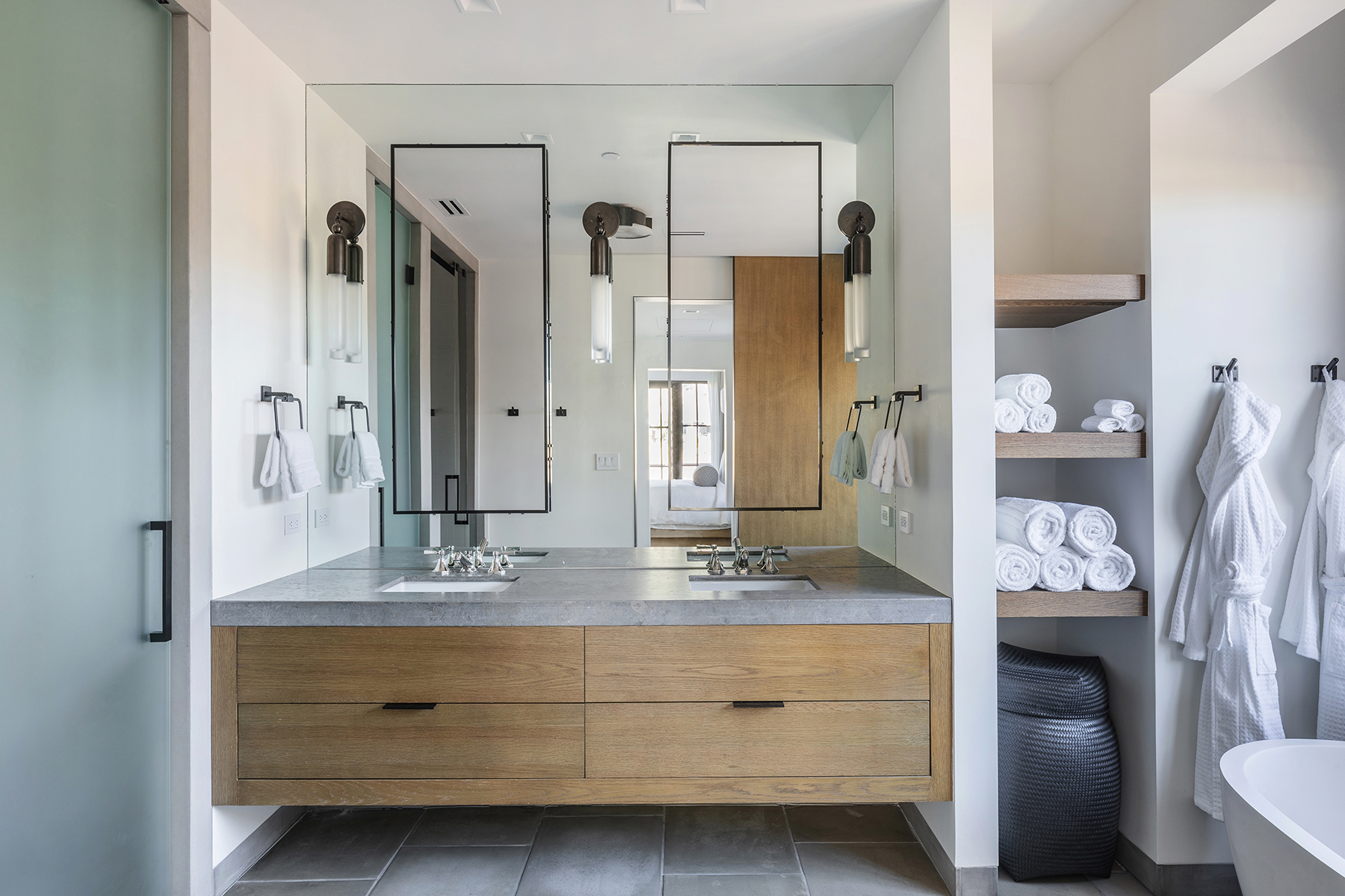 This modern primary bathroom features a sleek double-sink vanity with a grey stone countertop and light oak cabinetry. The space is accented by minimalist wall sconces, large floating mirrors, and open shelving stocked with neatly rolled white towels. A side area includes hanging robes, contributing to a clean, luxurious, and spa-like atmosphere.