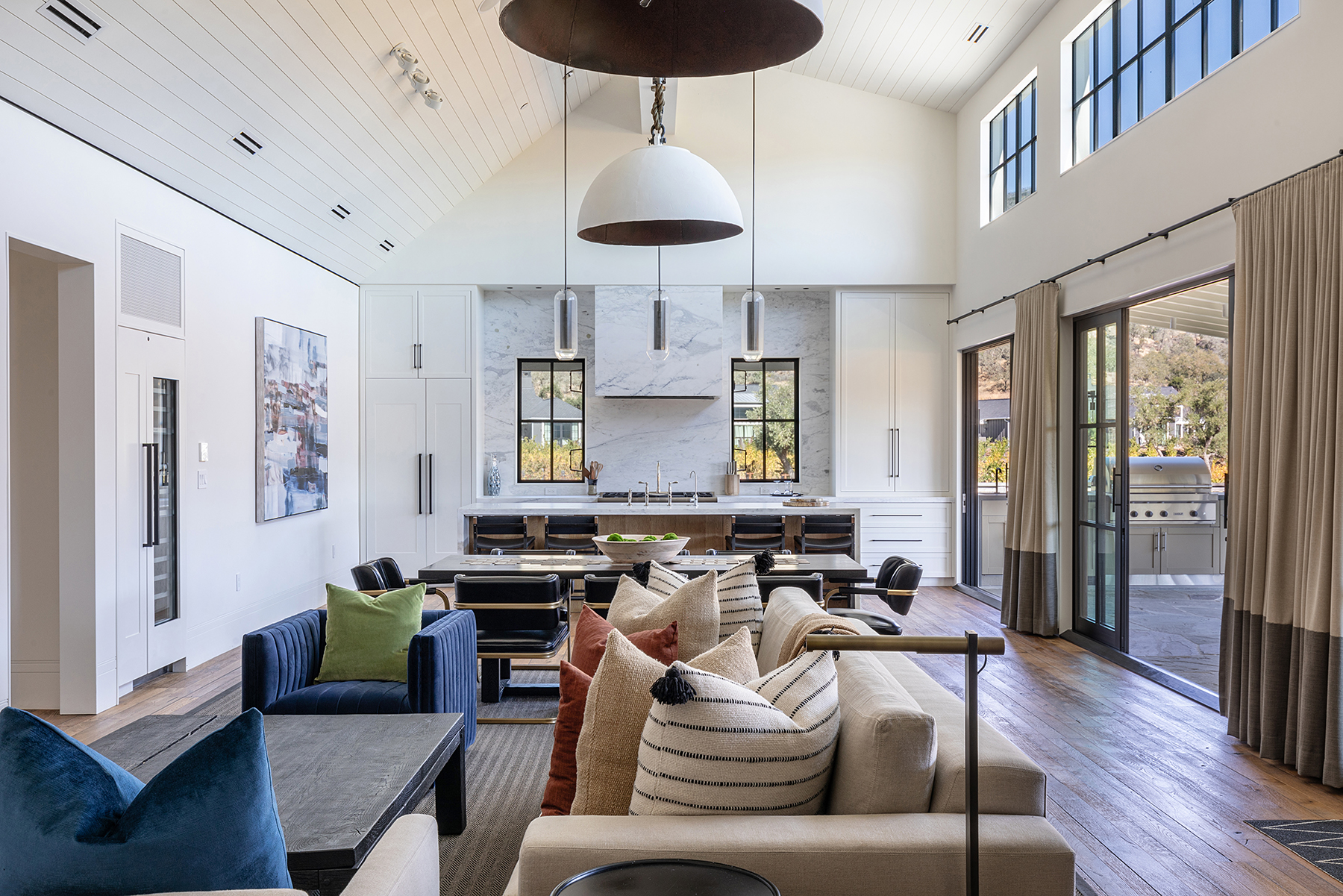 This spacious open-concept living area seamlessly integrates a cozy seating space with a sophisticated kitchen and dining room under a dramatic vaulted ceiling. The room features high-end finishes, including hardwood floors, a marbled backsplash in the kitchen, and stylish contemporary furniture, creating a warm and luxurious atmosphere. Large glass sliding doors connect the space to an outdoor patio, providing abundant natural light and ideal indoor-outdoor flow.