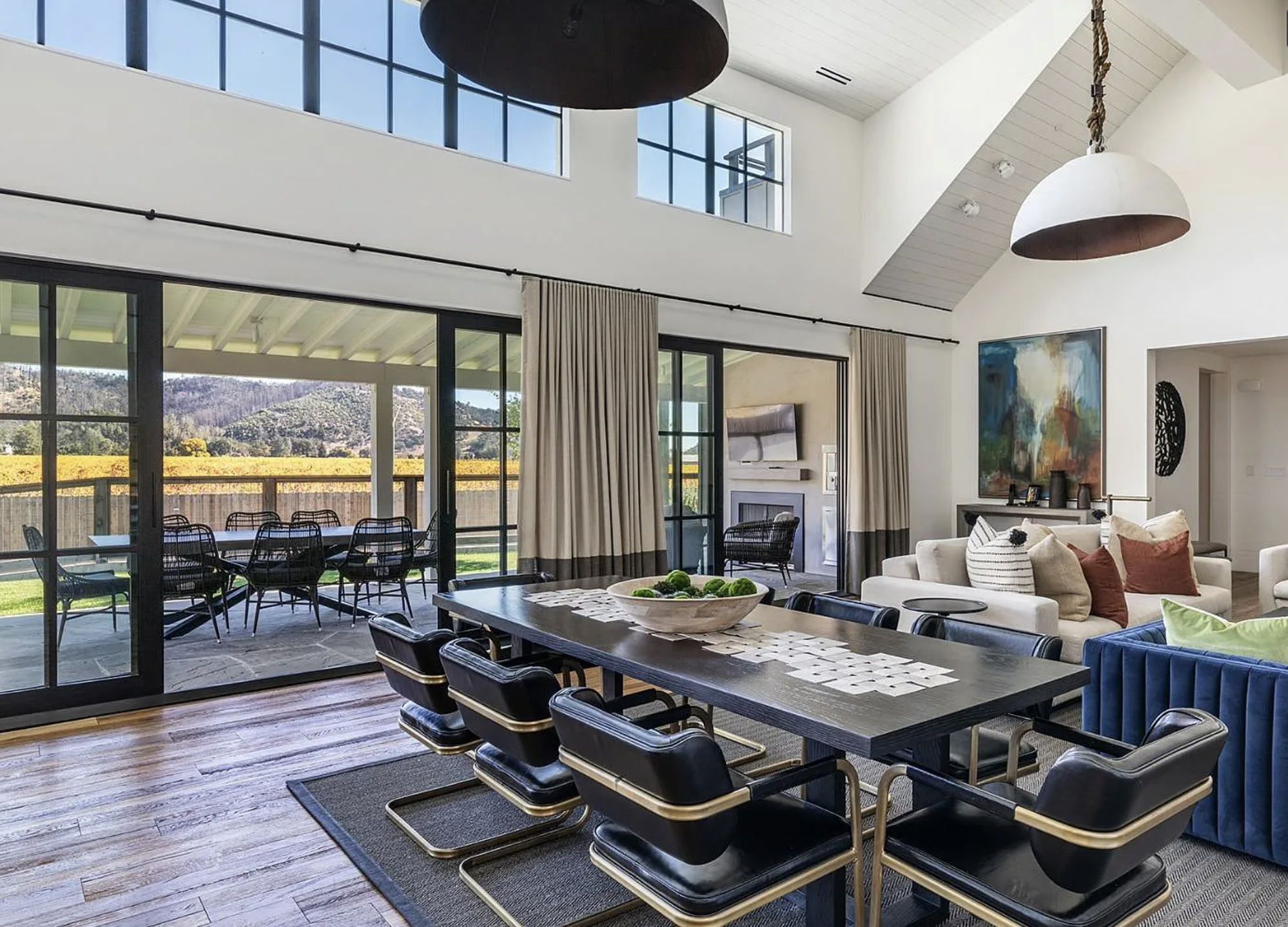 This elegant open-concept dining room features a long, dark wood table surrounded by modern leather and gold-accented chairs, set atop a textured area rug. With vaulted ceilings, transom-style windows, and seamless sliding glass doors that open to a scenic patio, the space feels bright, expansive, and perfectly integrated with the outdoors. The room's sophisticated decor, balanced by warm wooden flooring, creates an ideal setting for both casual meals and refined entertaining.