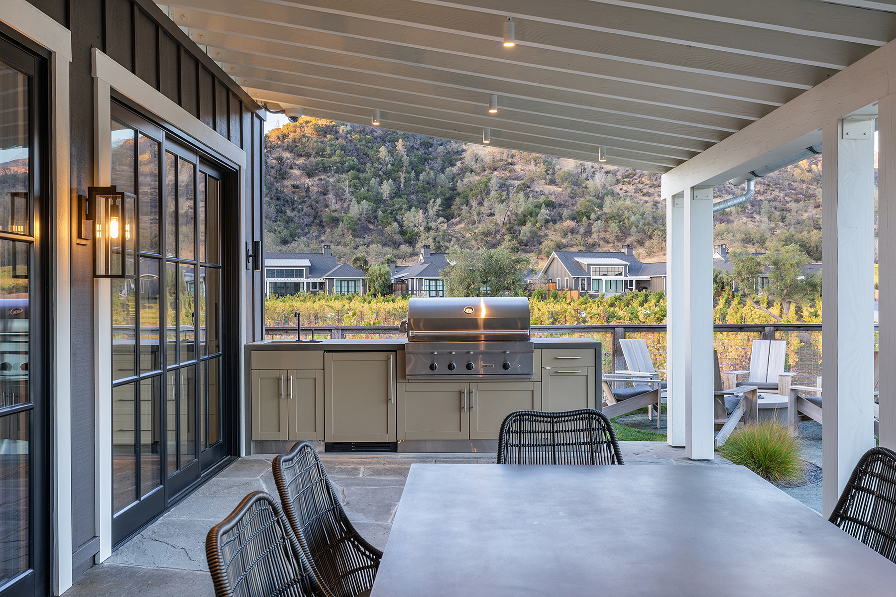 This covered outdoor patio features a sleek built-in grill station with high-end stainless steel appliances and muted-tone cabinetry, set against a backdrop of rolling vineyard hills. The space is furnished with contemporary wicker chairs and a large table, creating an elegant area for alfresco dining and entertaining. The stone flooring and modern lighting design contribute to a refined, indoor-outdoor living atmosphere.