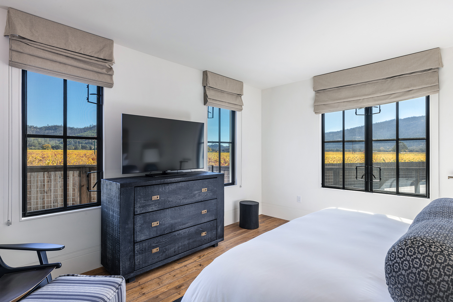 This primary bedroom features a modern, clean design with stark white walls and warm wood flooring. It includes a dark-stained dresser that holds a flat-screen television, flanked by large windows that offer views of the surrounding landscape. The space is well-lit and airy, with minimalist furniture that provides a peaceful and elevated aesthetic.