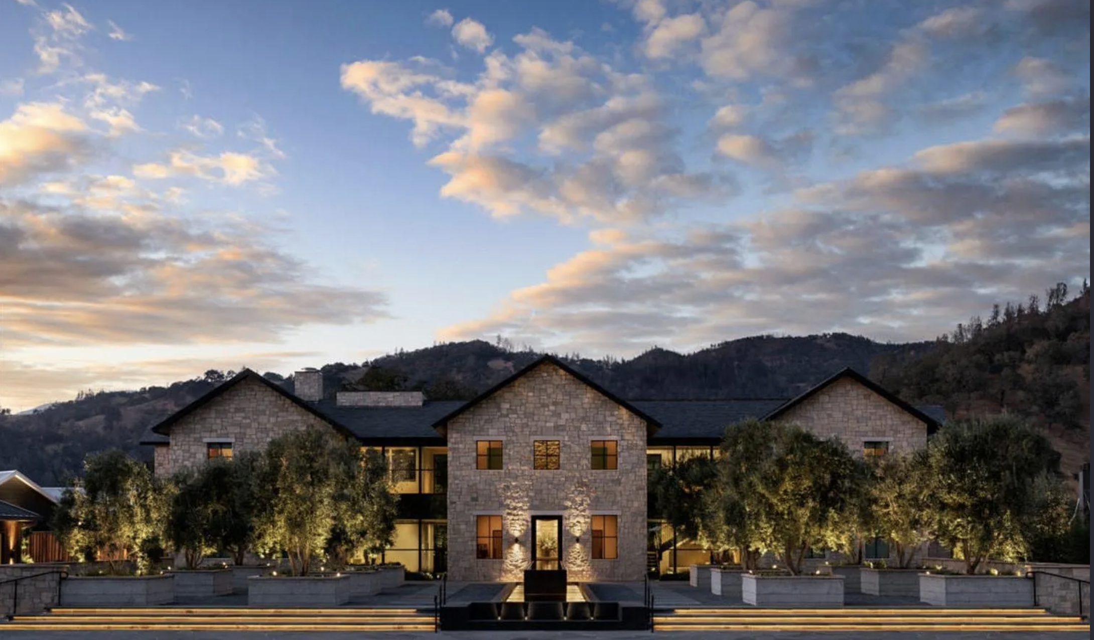 This stunning stone-clad residence showcases an elegant architectural design perfect for luxury living. The symmetrical facade is enhanced by warm exterior lighting, manicured trees in stone planters, and a sophisticated terraced entry with a water feature, creating an inviting and grand first impression.