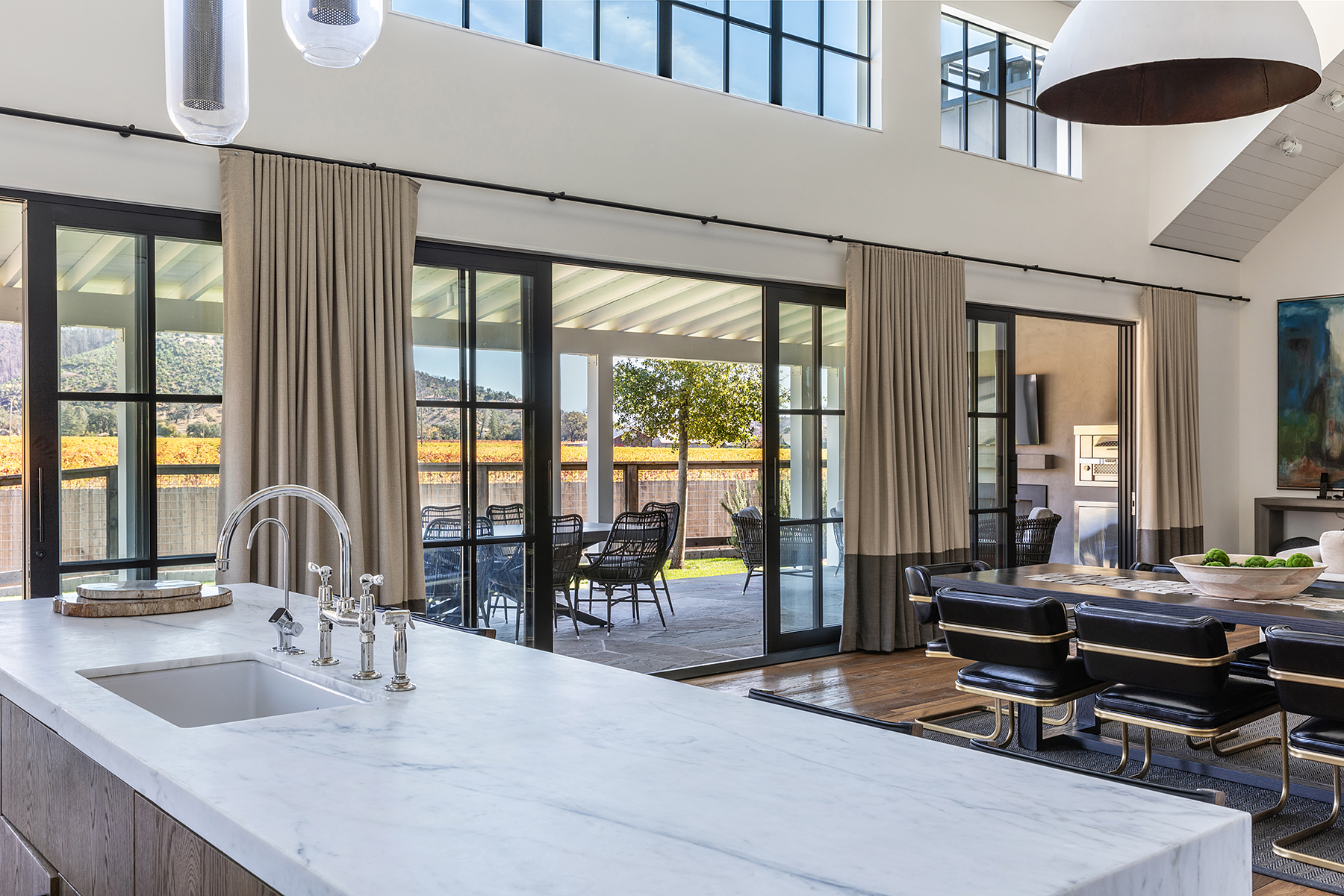 This stunning open-concept kitchen features a large marble-topped island with high-end polished chrome fixtures, seamlessly transitioning into an elegant dining area. Large floor-to-ceiling sliding glass doors provide a direct connection to a beautiful covered patio and offer expansive views of the picturesque vineyards outside. Its sophisticated design, high ceilings, and luxury finishes create a bright, airy, and inviting space perfect for modern entertaining.