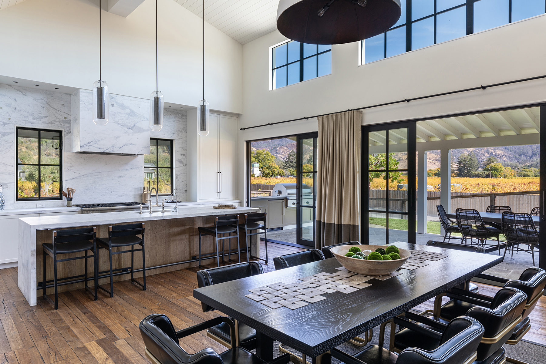 This spacious, open-concept kitchen and dining area features sophisticated modern design with vaulted ceilings, natural wood flooring, and elegant marble surfaces. Key highlights include a large kitchen island with bar seating, custom cabinetry, artistic pendant lighting, and seamless indoor-outdoor flow provided by expansive glass sliding doors. The room offers a stunning, sunlit atmosphere with picturesque views of the surrounding vineyard and rolling hills.