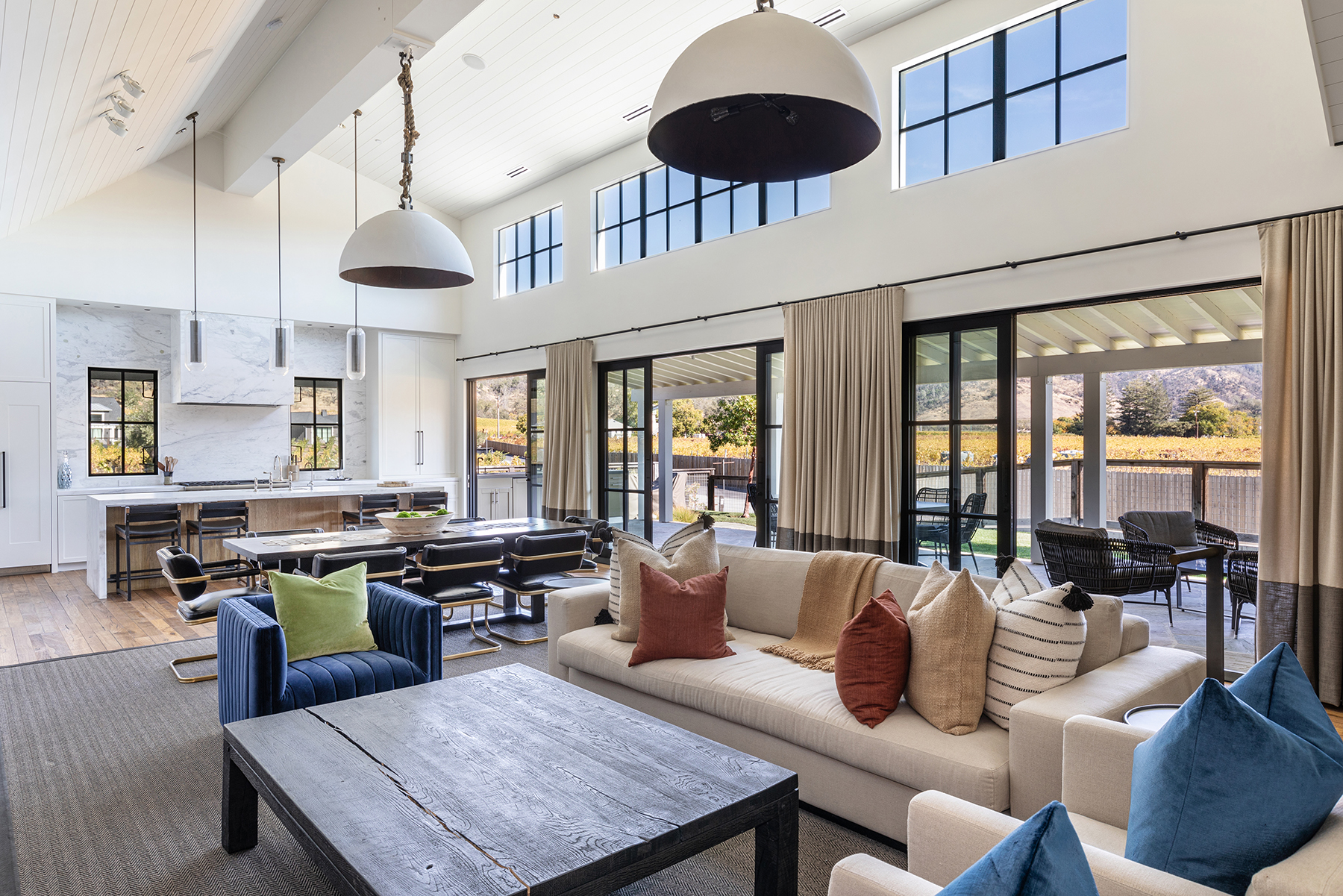 This spacious, open-concept living area features a modern, high-end design with vaulted ceilings and large glass sliding doors that seamlessly connect to an outdoor patio with vineyard views. The room is anchored by a large, light-colored sofa with accent pillows, a dark wooden coffee table, and a plush blue armchair, all highlighted by statement oversized pendant lights. The home exudes a luxurious, airy atmosphere, complemented by a gourmet kitchen in the background with marble countertops and a large dining island.