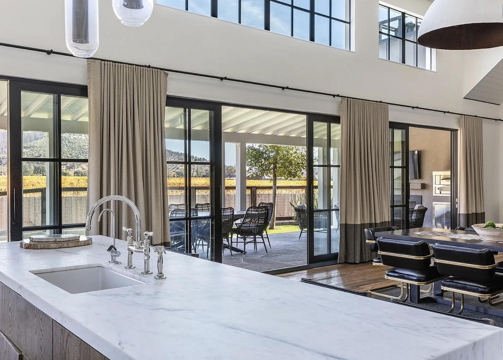 This sophisticated kitchen features a large, crisp marble-topped island with a sleek faucet, serving as a primary focal point. Expansive black-framed sliding glass doors open onto a covered patio, seamlessly blending indoor living with outdoor entertaining space while showcasing wine country vistas. The design exudes a modern, luxurious style with high ceilings, pendant lighting, and an adjacent dining area, creating an airy and open-concept atmosphere.