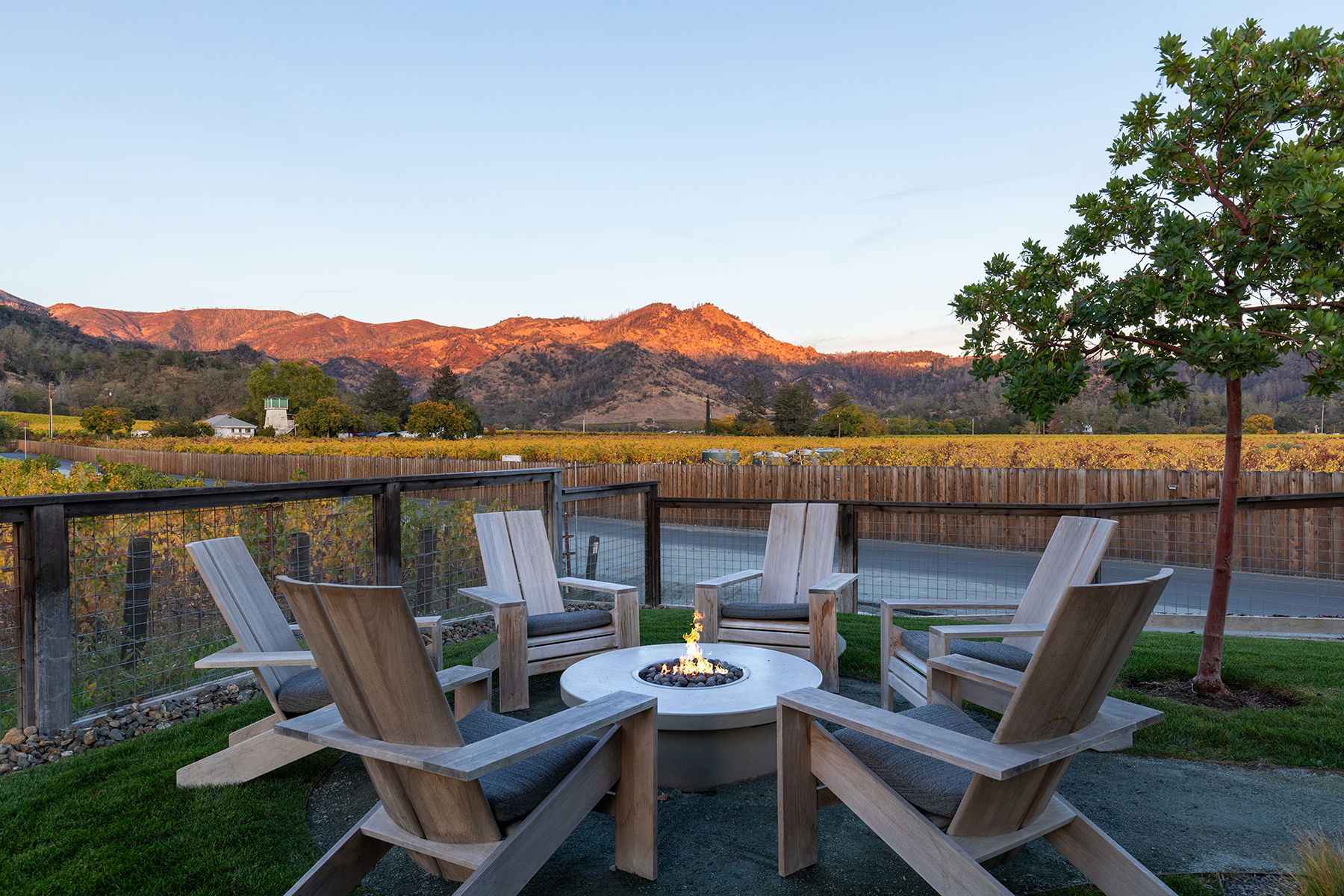 This inviting outdoor seating area features a modern circular concrete fire pit surrounded by five sleek, modern teak armchairs with grey cushions. The patio is set against a backdrop of a lush vineyard, a wooden perimeter fence, and stunning, sun-drenched mountain views at sunset. The space offers a sophisticated, peaceful atmosphere perfect for relaxation and capturing the beauty of a rural, natural landscape.
