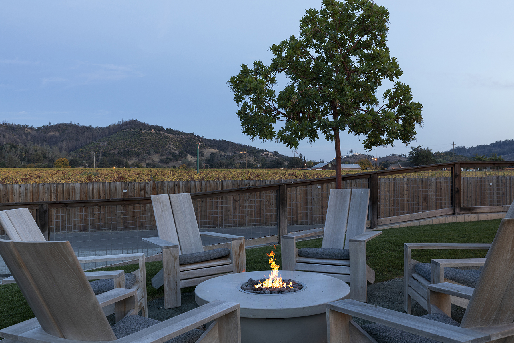 This outdoor space features a modern circular concrete fire pit surrounded by four comfortable lounge chairs, creating an inviting area for evening relaxation. The setting overlooks a scenic vineyard with soft hills in the background, offering a serene and picturesque landscape. It is an ideal spot for enjoying sunsets and outdoor entertaining within a peaceful, natural environment.