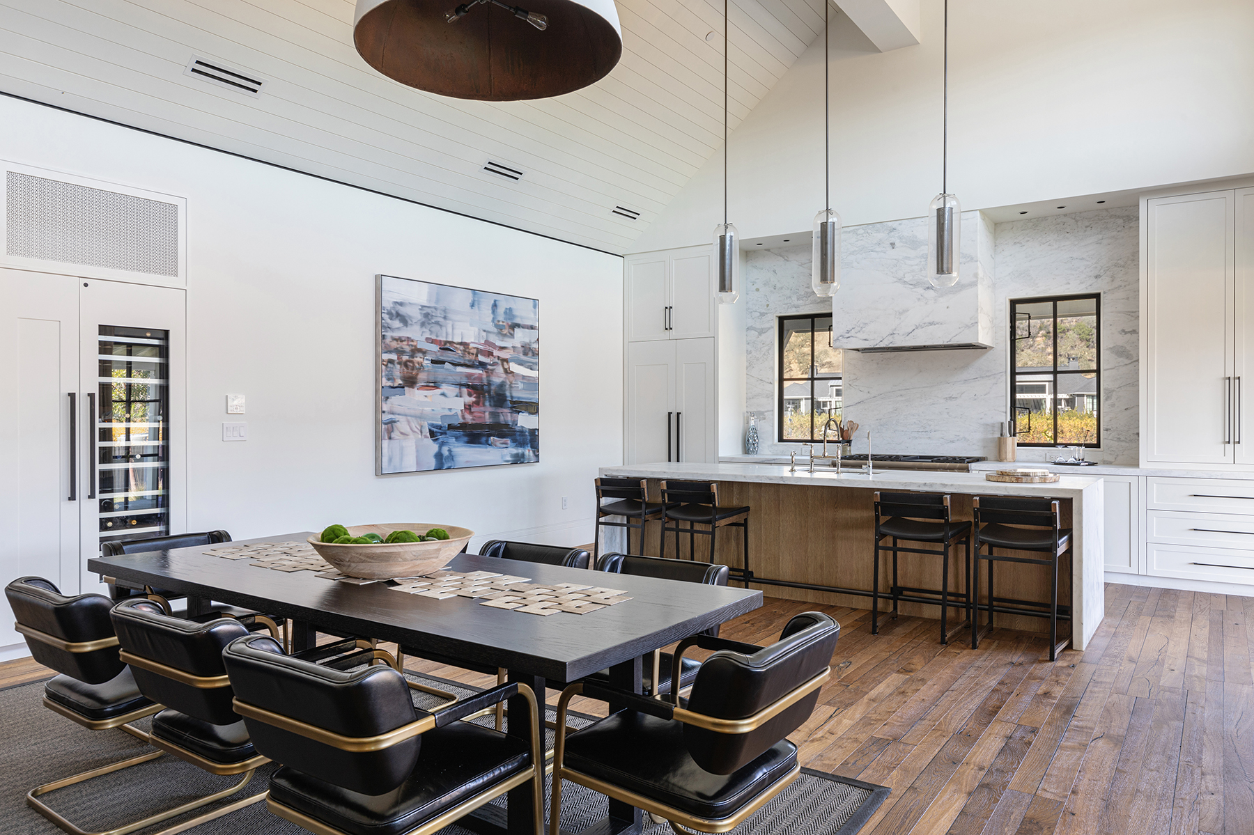 This stunning open-concept kitchen and dining area features a sophisticated blend of modern design and rustic textures, including a dark wood dining table paired with black and gold leather chairs. The kitchen boasts marble-topped counters, a waterfall edge island, and minimalist glass pendant lighting beneath a vaulted plank ceiling. Large windows bring in natural light, creating an airy, luxurious, and inviting atmosphere perfect for both casual dining and high-end entertaining.