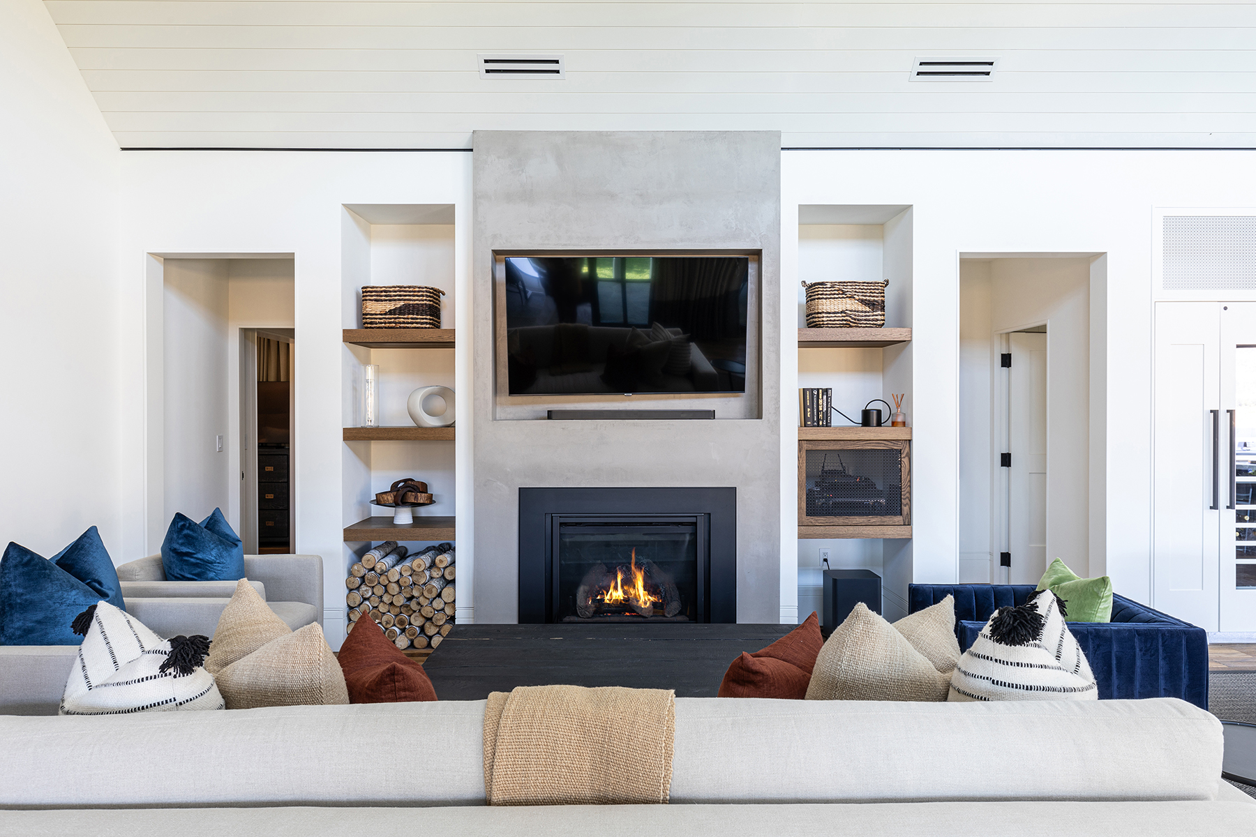 This sophisticated living room features a central fireplace with a concrete surround as the focal point, flanked by custom wood shelving units. The space is furnished with plush velvet and neutral-toned seating, highlighted by decorative pillows, creating a warm and inviting atmosphere. The design seamlessly blends modern architecture with cozy, textured elements, perfect for comfortable family living and entertaining.