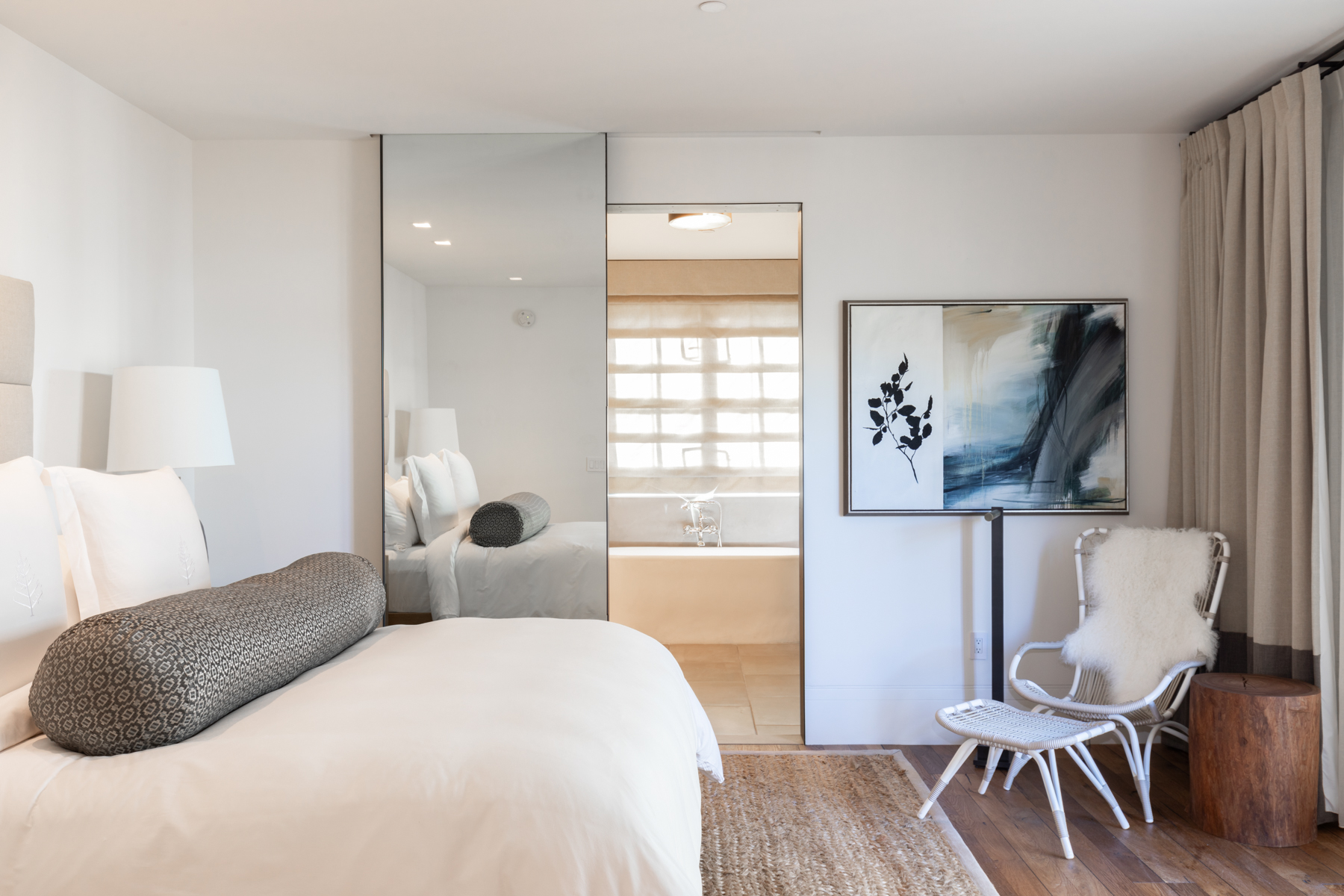 This elegantly appointed primary bedroom features a clean, minimalist aesthetic with neutral tones and natural textures, including a woven rug and a wooden side table. A seamless full-length mirror enhances the sense of space and provides a reflective view into the ensuite bathroom, which is equipped with a soaking tub. The room is completed by a contemporary armchair with a faux-fur throw and sophisticated abstract artwork, creating a serene and luxurious retreat.