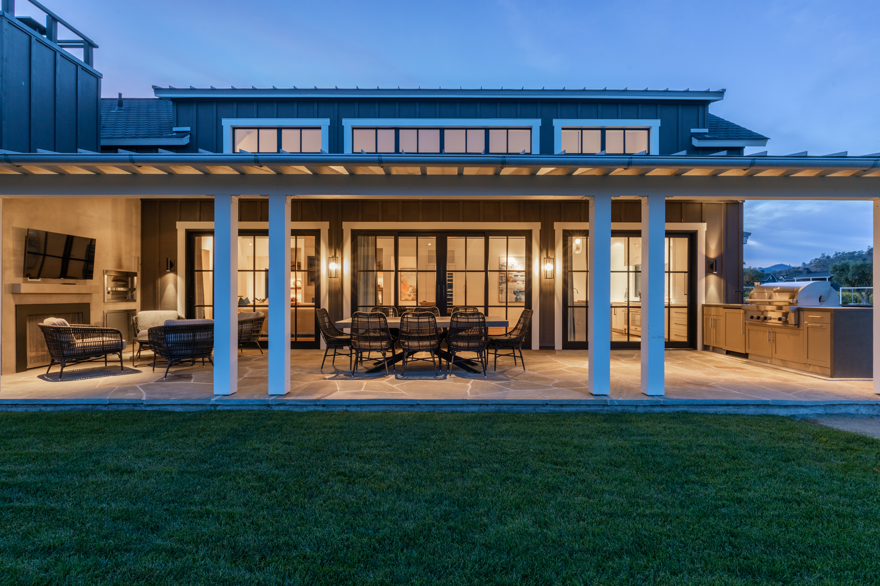 This modern farmhouse-style rear patio features a spacious stone-paved floor, a built-in outdoor kitchen, and a cozy lounge area with a fireplace and television. The space is covered by a structural beam roof and opens directly onto a manicured grass yard through elegant black-framed glass sliding doors. Its thoughtful design creates a seamless indoor-outdoor living experience, ideal for evening entertainment.