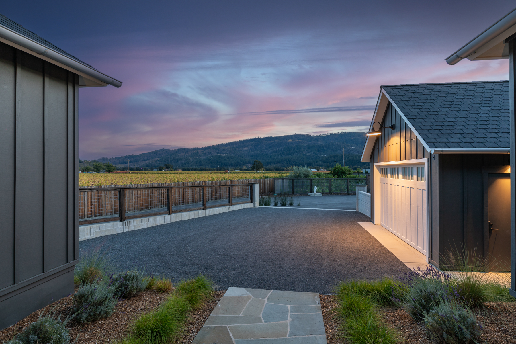 This image features a modern, dark-sided residence focusing on the attached garage driveway area at dusk. A scenic view of rolling hills and vineyards is visible in the background, complemented by artistic landscaping and a welcoming glowing exterior light. The clean lines and contemporary farmhouse aesthetic create an inviting and peaceful atmosphere.
