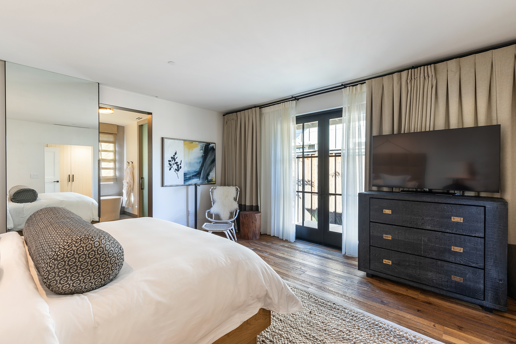 This sophisticated primary bedroom features a plush king-sized bed with a patterned bolster pillow and a sleek dark wood dresser topped with a flat-screen television. The space is accented by wide-plank hardwood floors, a textured area rug, and an adjoining spa-like bathroom visible through a glass partition. Large windows dressed in elegant floor-to-ceiling drapes invite natural light, creating a serene and luxurious retreat.