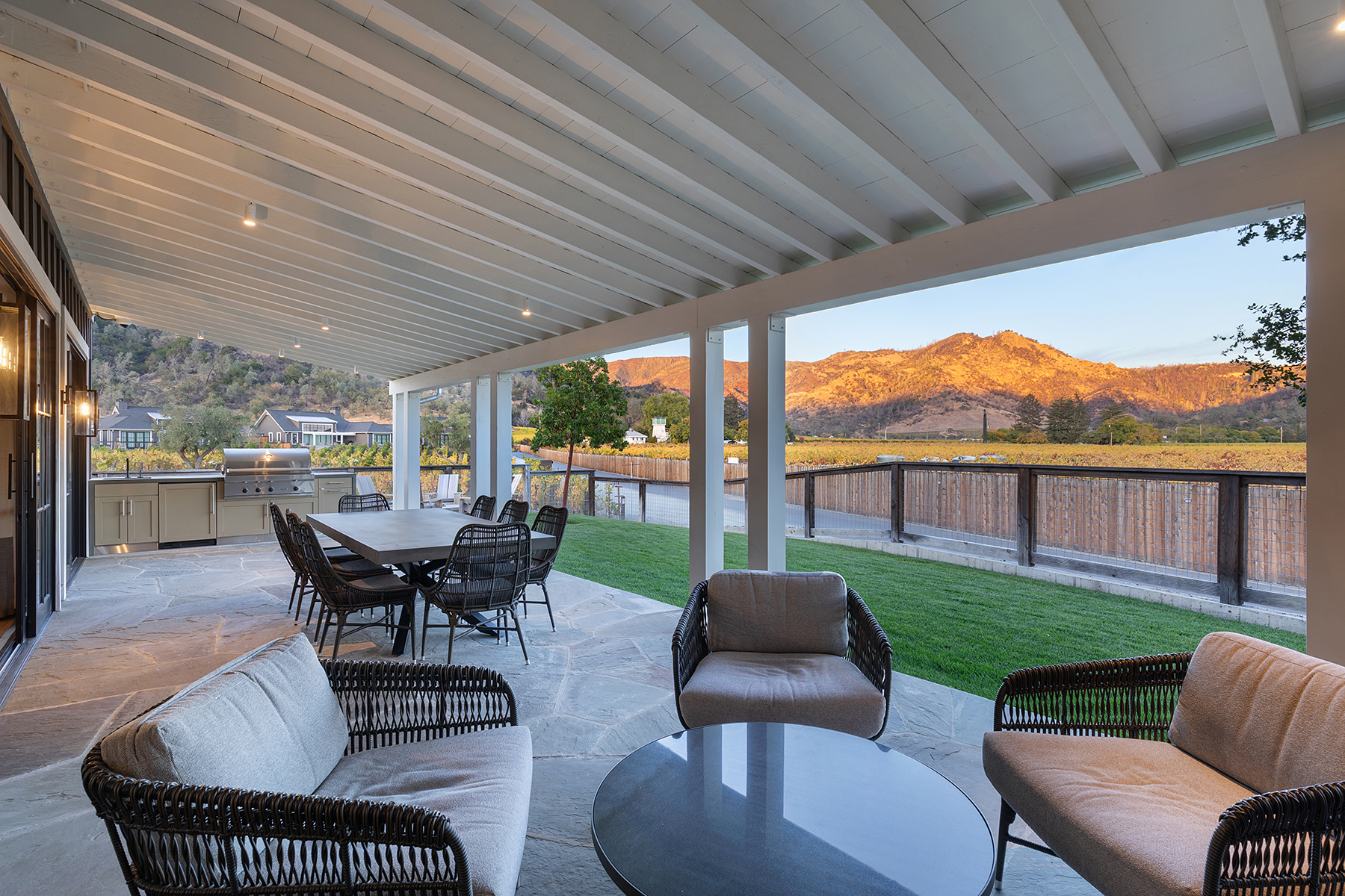 This luxurious covered patio features stone slab flooring, comfortable outdoor lounge furniture, and a dedicated dining area adjacent to an outdoor kitchenette. The space seamlessly bridges the indoor living area with a beautifully manicured lawn, offering stunning, unobstructed views of the sunset-lit hills and the surrounding vineyard landscape.