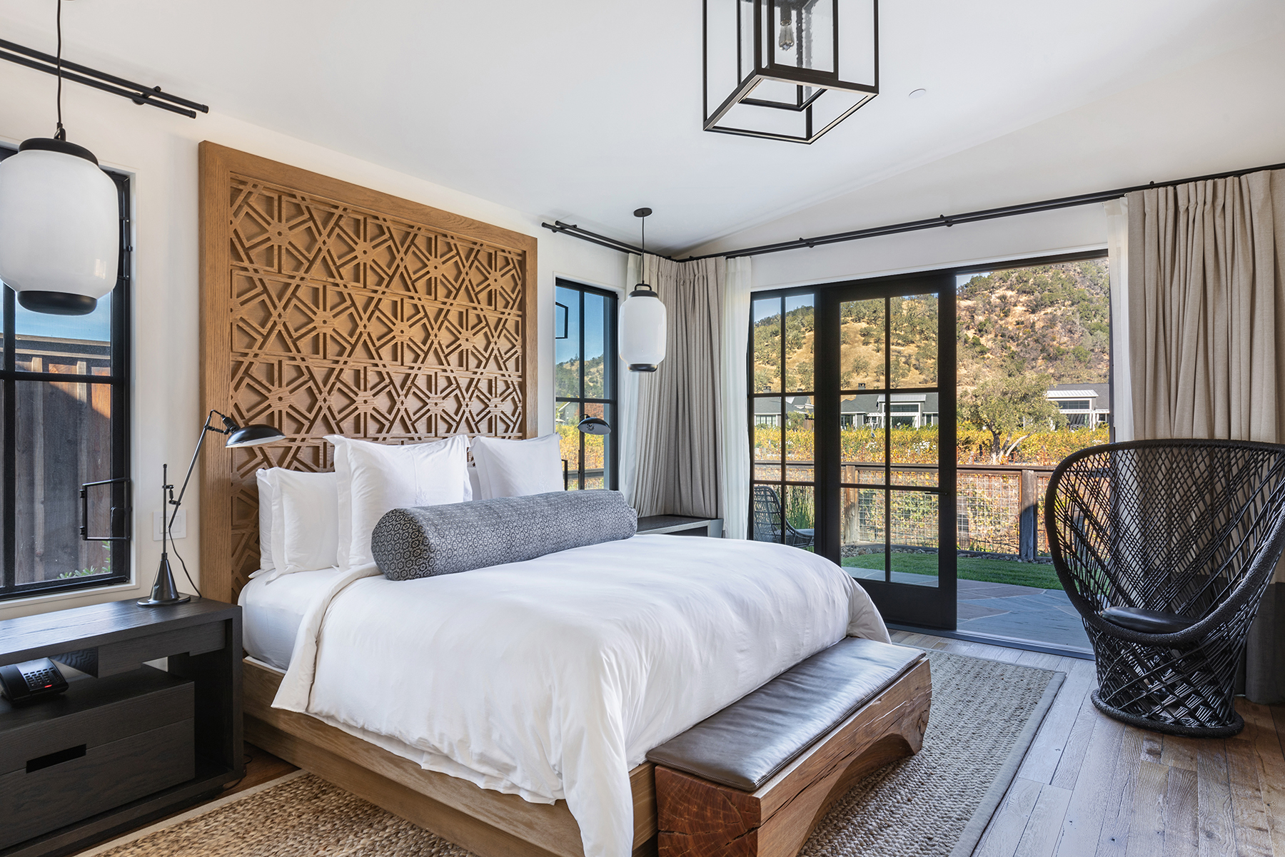 This sophisticated primary bedroom features a stunning custom wood geometric headboard as its focal point, paired with neutral bedding and a textured bolster pillow. Large floor-to-ceiling glass doors provide abundant natural light and seamless access to a private outdoor patio overlooking a landscape, enhancing the serene, upscale aesthetic. The room is completed by a dark wood bedside table, a modern black wicker chair, and elegant light fixtures, creating a refined and tranquil retreat.