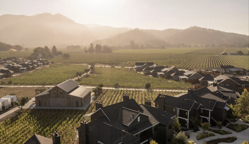 This aerial view captures a luxury residential community seamlessly integrated with lush, expansive vineyards. The scene features modern, dark-toned homes and a rustic wooden barn, set against a backdrop of rolling, hazy hills under a warm, golden sunset. The overall atmosphere is one of sophisticated, rural elegance and tranquility.