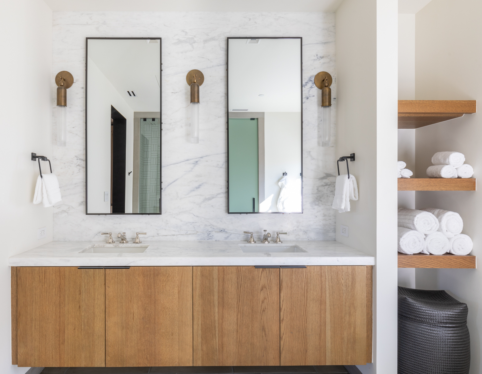 This sleek and modern primary bathroom features a double vanity with a warm wood cabinet base topped by a pristine white marble countertop and matching marble backsplash. The space is highlighted by two vertical rectangular mirrors, elegant brass sconce lighting, and open wooden shelving on the side containing neatly stacked towels. The overall aesthetic is clean, luxurious, and highly sophisticated.