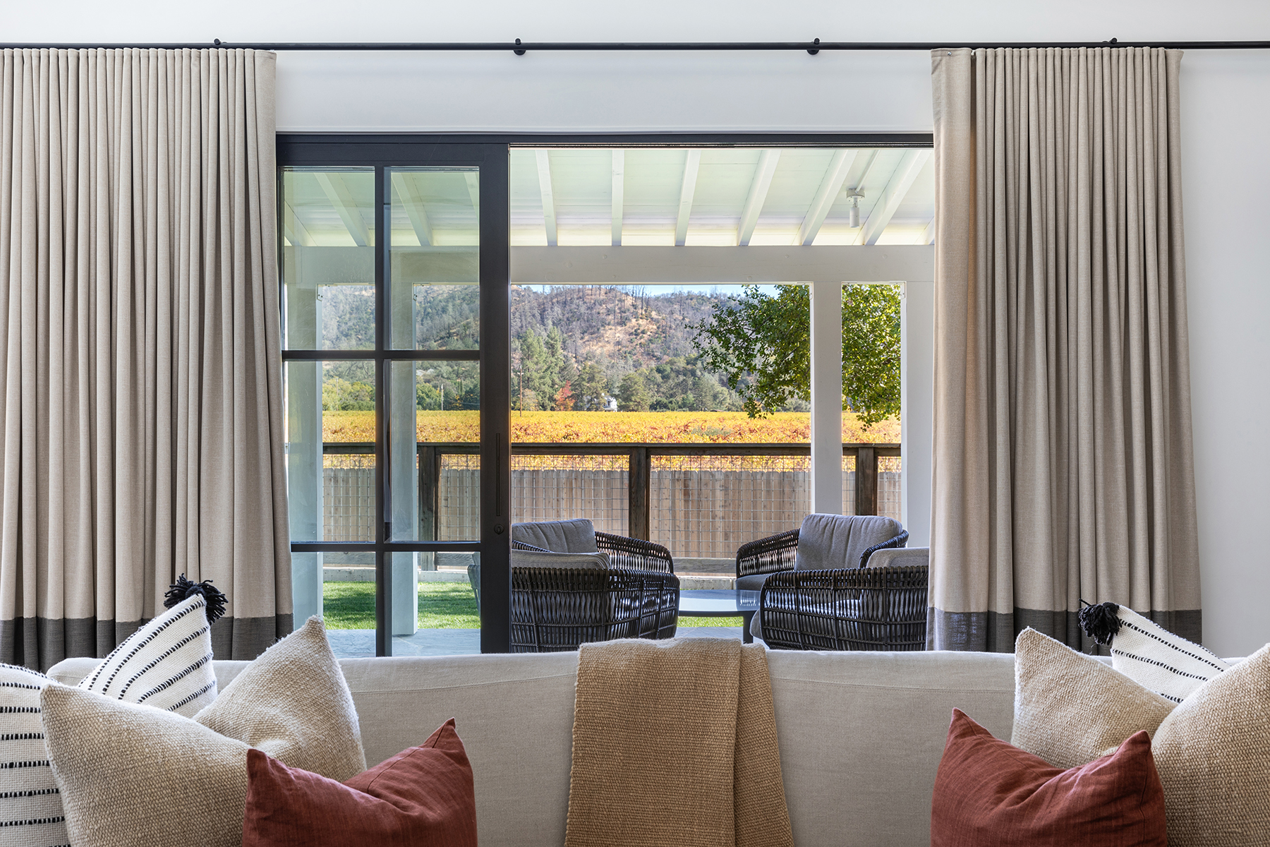 This interior view showcases a comfortable, transitional-style living room centered around a plush neutral sofa accented with textured throw pillows. Large floor-to-ceiling glass sliding doors lead to an inviting covered patio, offering stunning views of a vineyard and distant hills. The room feels bright, serene, and seamlessly connected to the outdoor landscape.