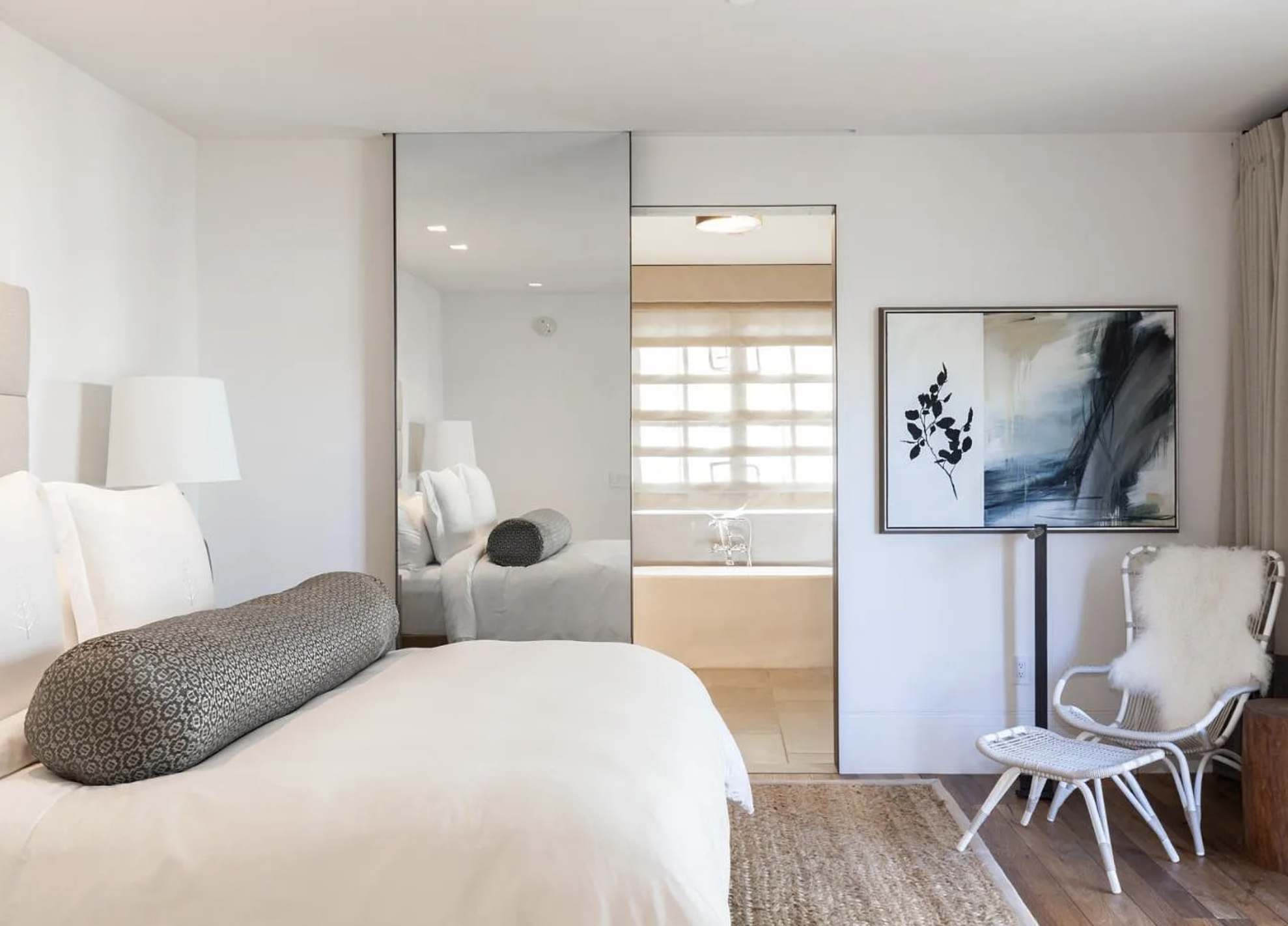 This serene and modern primary bedroom features a plush white bedding set accented by a patterned bolster pillow and crisp white walls. A large floor-to-ceiling mirror adds depth to the room while offering a framed view into the adjacent soaking tub in the bathroom. The space is softened by a natural jute area rug and a designer white chair with a faux fur throw, creating a balanced and inviting atmosphere.