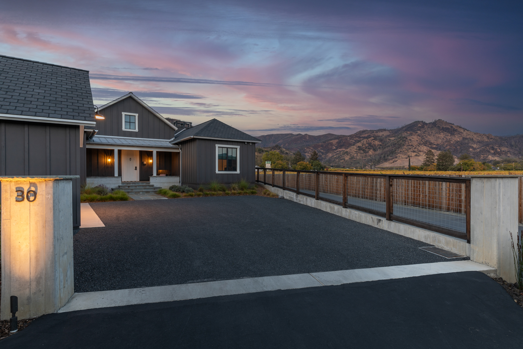 This modern farmhouse-style home features dark vertical siding, crisp white trim, and a welcoming entryway. The property includes a sleek, gravel-paved driveway, a contemporary concrete pillar with illuminated address numbering, and a rustic wood-and-wire fence overlooking a vineyard landscape at sunset. The architectural design combines clean, sophisticated lines with cozy, inviting lighting, suggesting an elegant and well-maintained private residence.