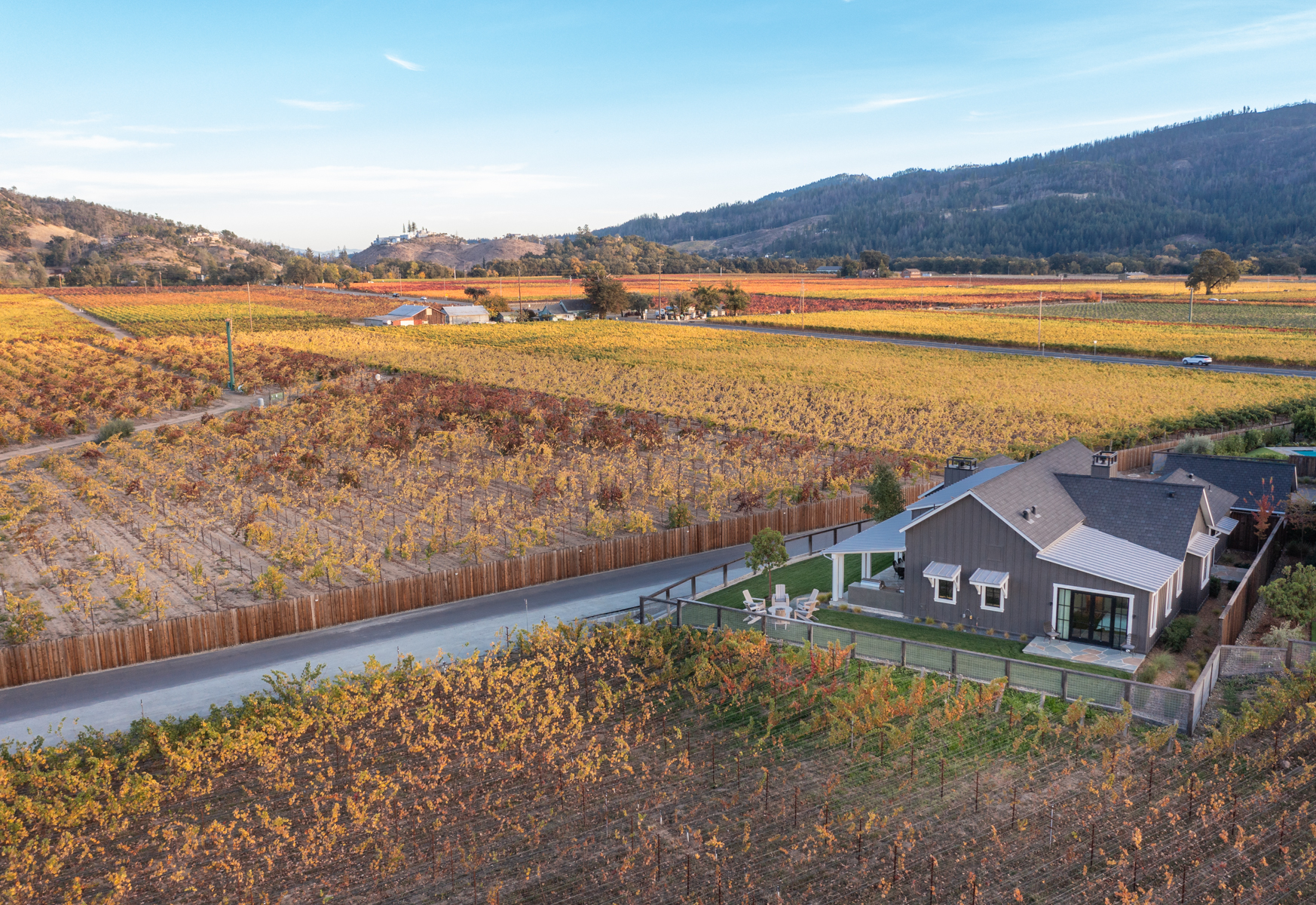 This aerial view showcases a modern dark-sided farmhouse situated amidst vast, golden-hued vineyards. The property is well-manicured, featuring a neat lawn, a cozy patio seating area, and a rustic fence line that integrates perfectly into the rural landscape, highlighting a serene and sophisticated vineyard estate lifestyle.