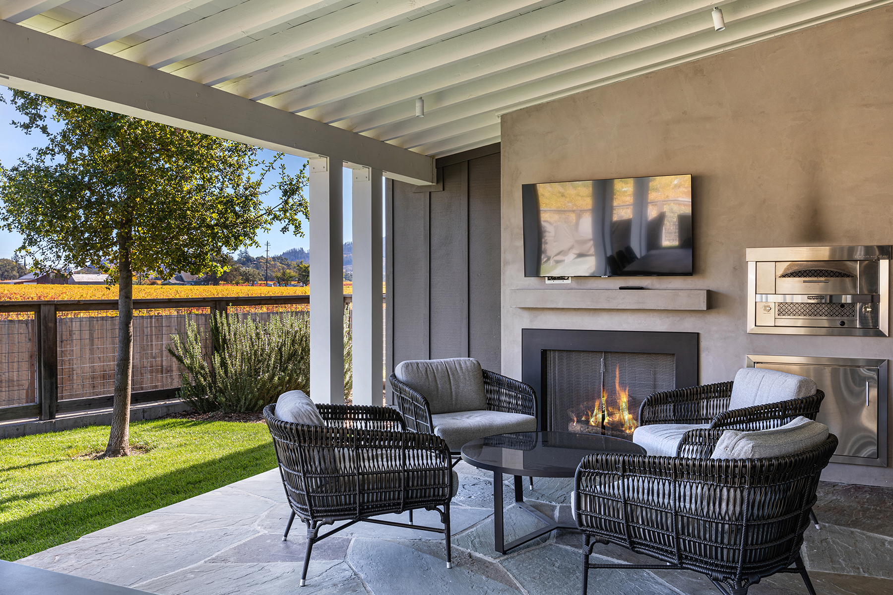 This covered outdoor patio area features a modern stone fireplace integrated into a textured wall, complete with a wall-mounted television above. The space is comfortably furnished with four wicker chairs surrounding a round table, all set on beautiful flagstone flooring. Offering a serene view of a lush green lawn and an adjacent vineyard, the patio provides a sophisticated and inviting setting for relaxation and outdoor entertaining.