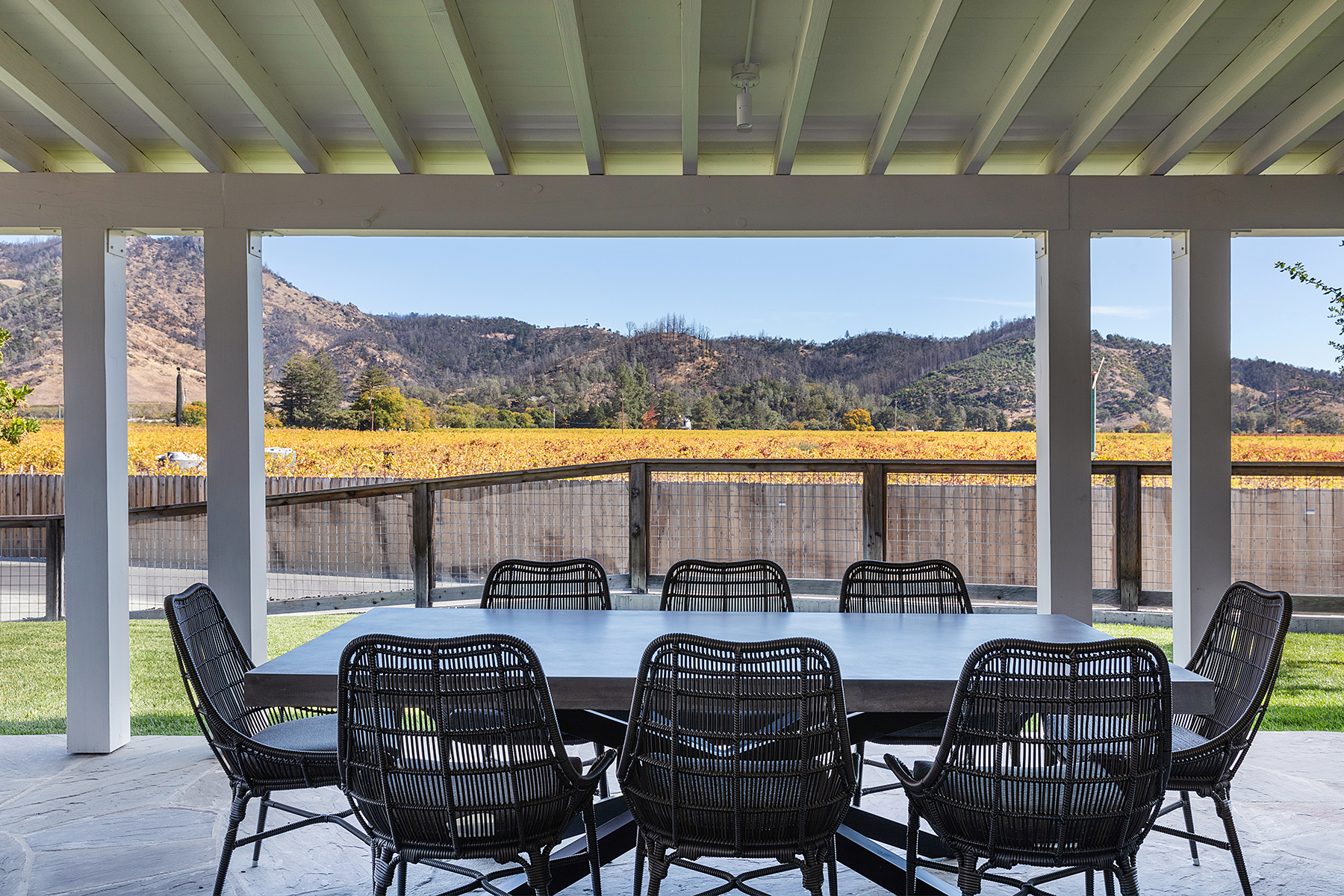 This covered outdoor patio features a large, modern rectangular dining table surrounded by eight stylish black wicker chairs. The space offers a picturesque, unobstructed view of golden-hued vineyards set against a backdrop of rolling hills. The architectural design includes an exposed beam ceiling, creating an inviting and well-composed transitions between indoor comfort and natural scenery.