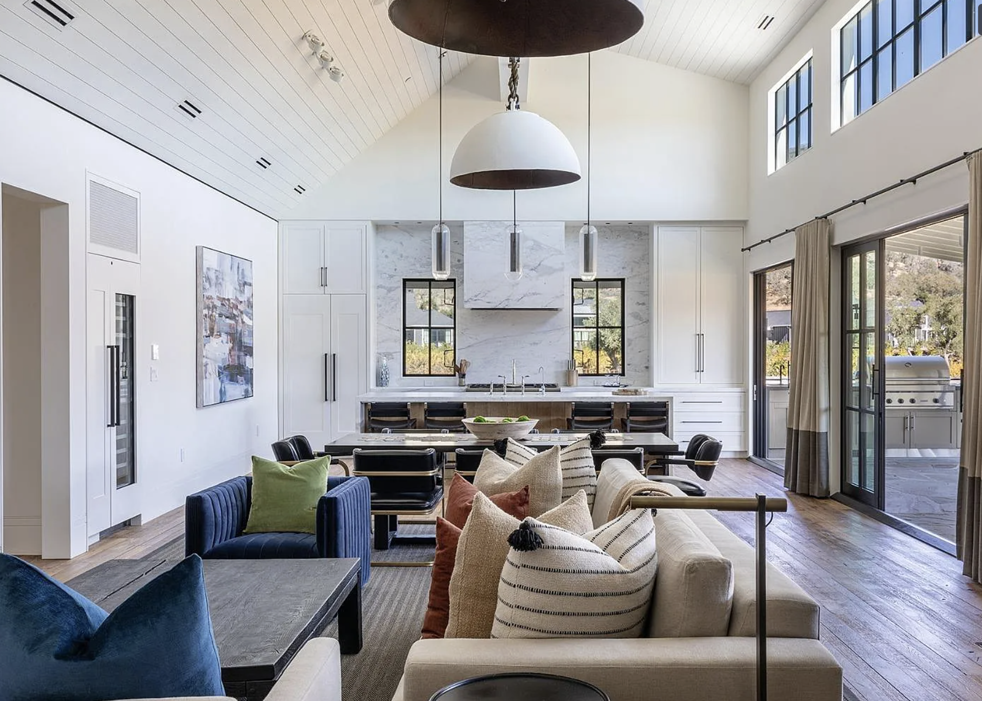 This spacious open-concept living area seamlessly transitions into a high-end kitchen, highlighting a vaulted, plank-style ceiling and warm wooden flooring. The space is anchored by a large, comfortable sofa and velvet armchairs, complemented by sophisticated neutral and earth-tone textiles. Expansive glass doors open to a rear patio, creating an airy, light-filled environment ideal for both relaxation and entertaining.