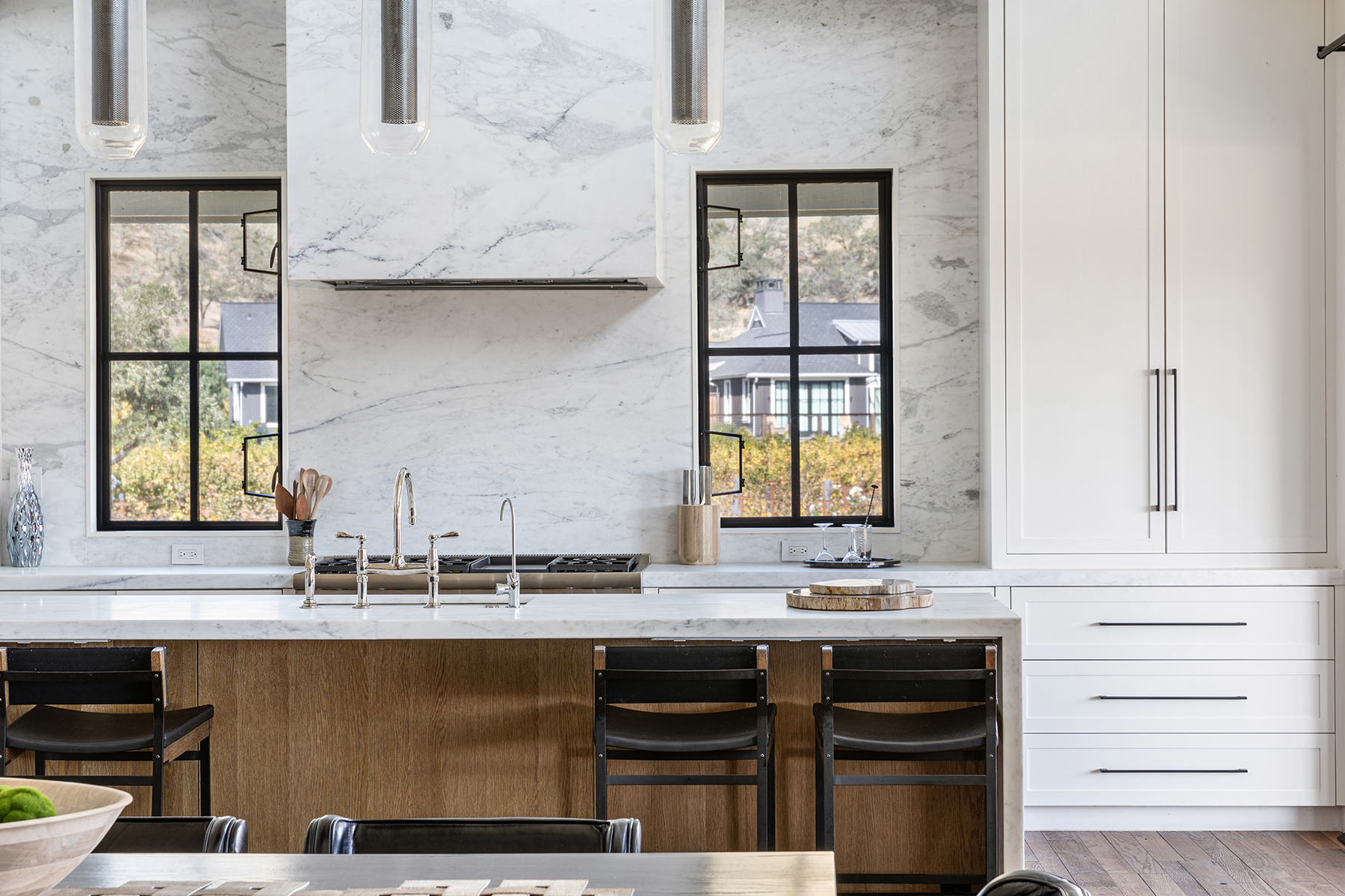 This sophisticated, modern kitchen features a spacious marble-topped island with warm wood cabinetry and black bar stools. A stunning marble waterfall-style hood anchors the cooking area, which is framed by symmetrically placed windows that provide natural light and picturesque views. The clean lines, premium fixtures, and high-quality materials create a bright, contemporary, and luxurious space perfect for both cooking and gathering.