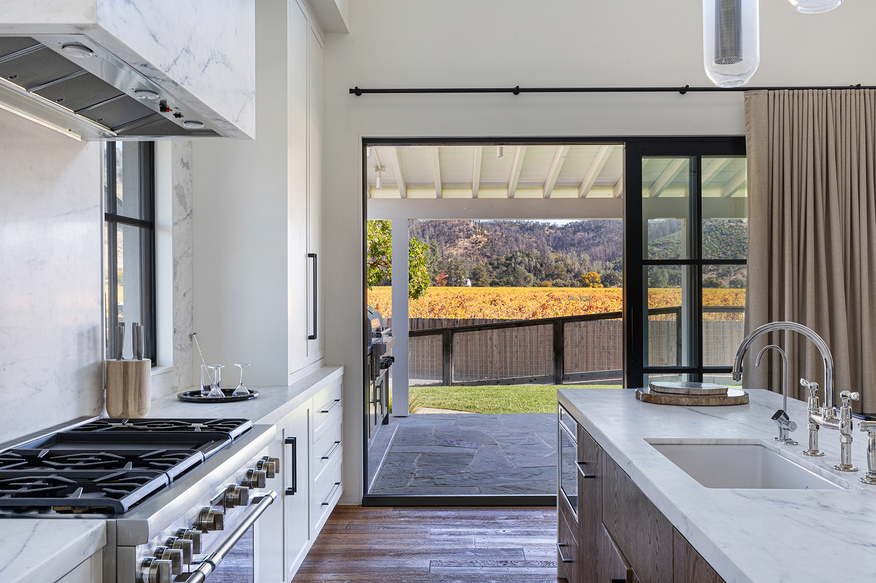 This sophisticated kitchen features a seamless blend of white marble countertops and dark wood cabinetry, creating a clean, high-end aesthetic. A large sliding glass door seamlessly connects the indoor cooking space to a stone-paved patio and a scenic vineyard view beyond. The room is equipped with professional-grade appliances, including a prominent range, making it both functional and visually stunning for a luxury home.