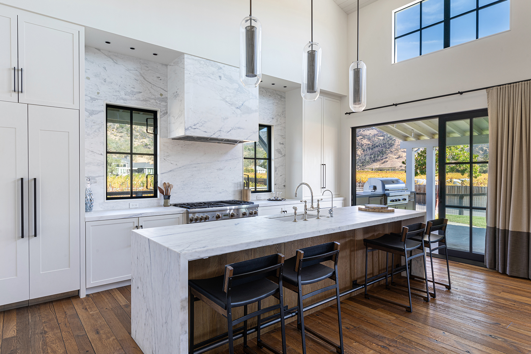 This stunning contemporary kitchen features high-end marble countertops, a seamless marble slab backsplash and hood, and custom white cabinetry. The space is anchored by a large kitchen island with leather-upholstered bar stools and warm wooden paneling, complemented by elegant pendant lighting. Large windows and glass sliding doors provide abundant natural light and picturesque views of the surrounding vineyard landscape.