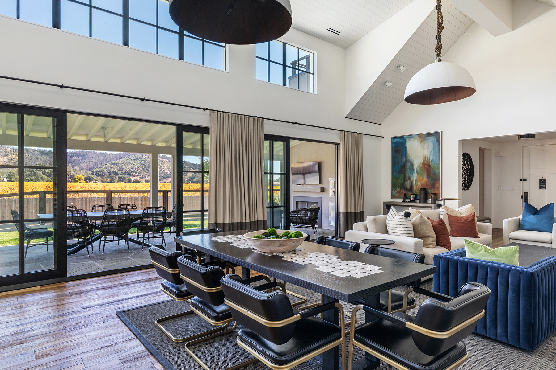 This elegant open-concept dining space features a sleek black table surrounded by modern, gold-accented leather chairs, positioned beneath a dramatic pendant light. The room is flooded with natural light from expansive floor-to-ceiling windows that offer sliding glass door access to a covered patio with vineyard views. The sophisticated design integrates seamlessly into a living area with contemporary furniture, reflecting a luxurious and airy architectural style.
