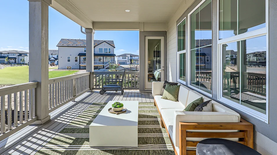 This image showcases a stylish outdoor patio area, featuring a modern white sofa with green pillows, a white coffee table with a potted plant, and a striped outdoor rug. The patio is partially covered, with views of a well-maintained lawn and neighboring houses in the background. Large windows provide ample natural light and a seamless transition between indoor and outdoor living spaces.