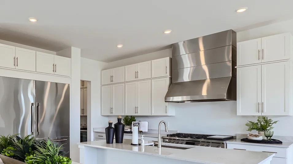This is a bright and modern kitchen featuring white cabinets, stainless steel appliances, and a large island with a white countertop. The stainless steel range hood stands out as a focal point, and the overall design is clean and contemporary. The perspective is from a medium distance, showcasing the kitchen's layout and key features.