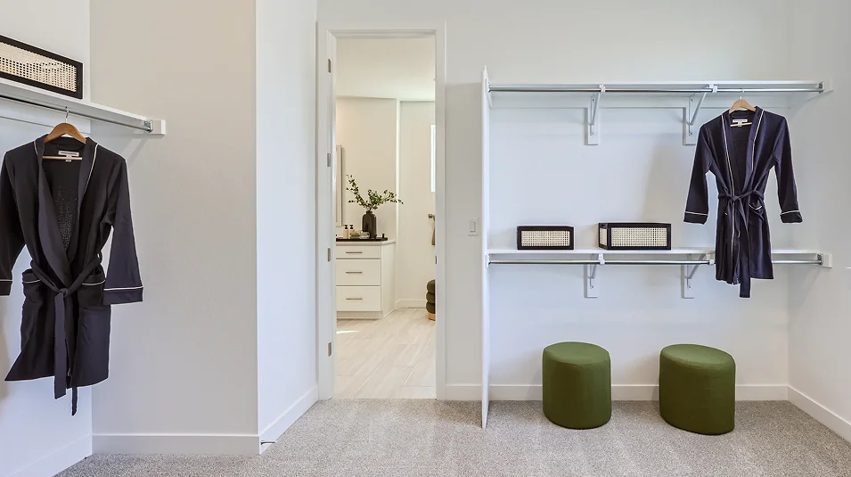 This is a bright and organized walk-in closet featuring a neutral color palette. The closet includes shelving with decorative boxes, hanging rods with robes, and two green ottomans. An open doorway leads to a bathroom, creating a seamless transition between spaces.