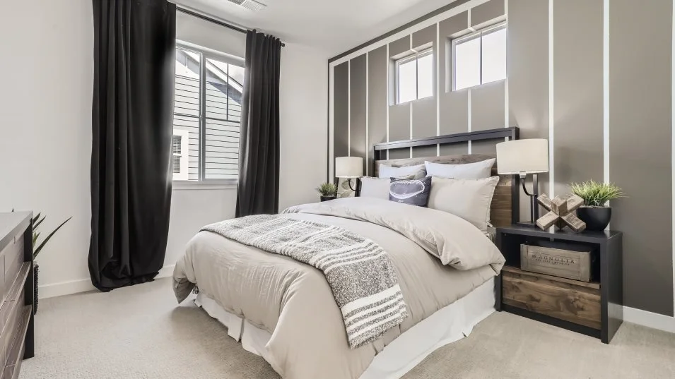 This is a well-staged primary bedroom featuring a neutral color palette with gray and white accents. The bed is neatly made with a beige duvet and a patterned throw blanket. The room has a modern design with a decorative wall behind the bed and dark curtains framing the window, creating a cozy and inviting atmosphere.