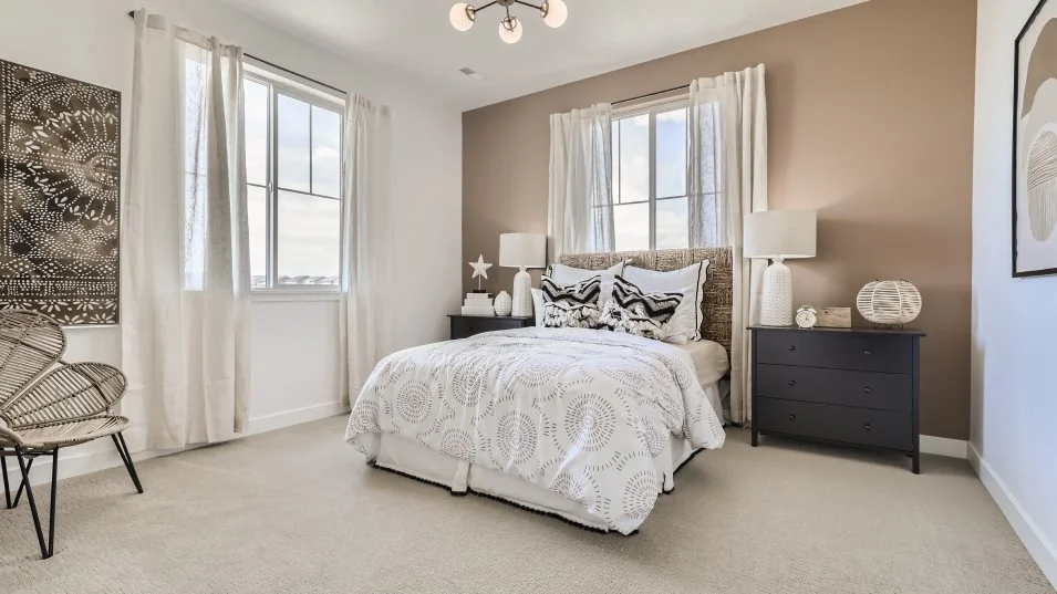 This is a well-lit guest bedroom featuring a neutral color palette with white walls and beige accents. The room includes a bed with patterned bedding, two windows with sheer white curtains, and a dark-colored nightstand with a lamp. A decorative chair and artwork add to the room's aesthetic, creating a comfortable and inviting space.