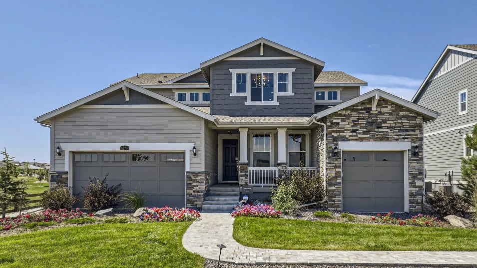 This is a front view of a two-story home with a well-manicured lawn and landscaping. The house features a combination of stone and siding, with a covered front porch and a two-car garage. A paved walkway leads to the front door, enhancing the curb appeal and creating an inviting entrance.